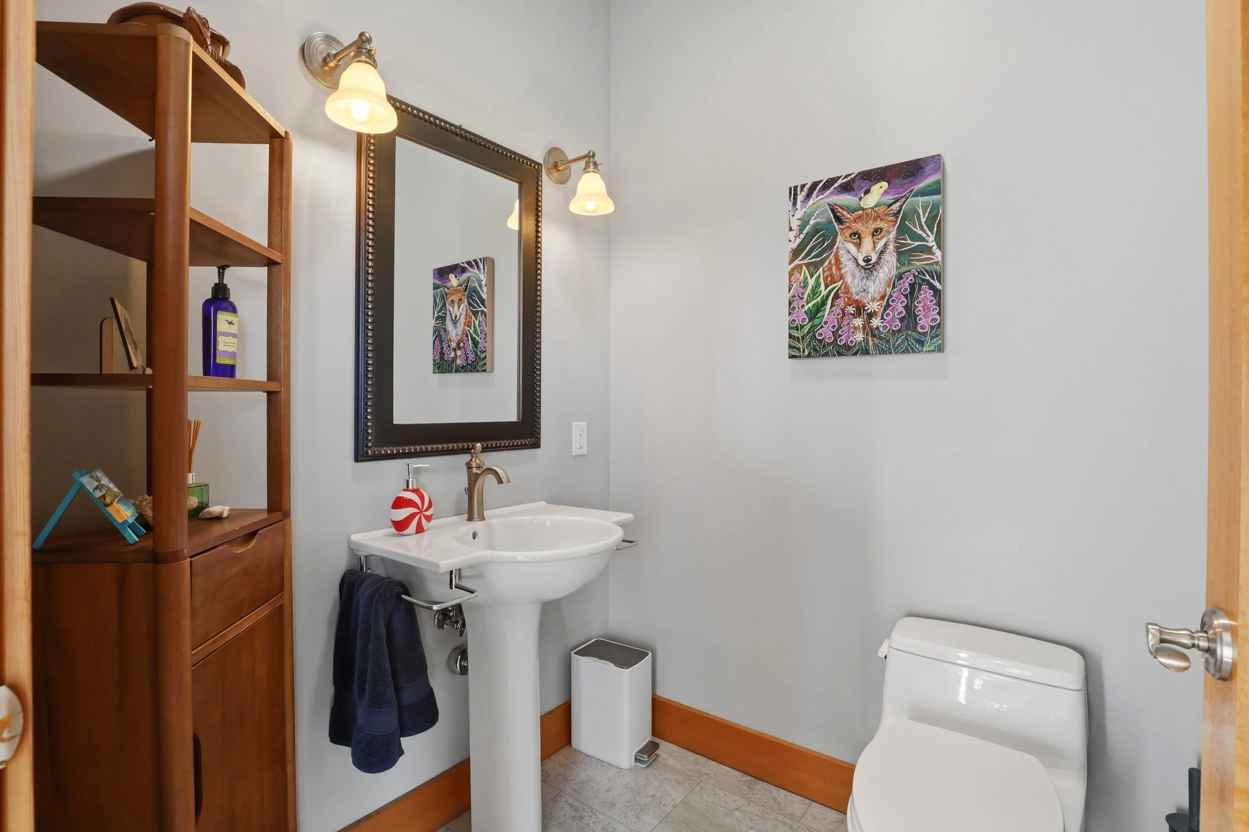 A small bathroom with a wooden shelf, a mirror with beaded frame, wall-mounted light fixtures, a painting of a fox with colorful foliage, a small sink with a striped soap dispenser, a toilet, and a trash bin.