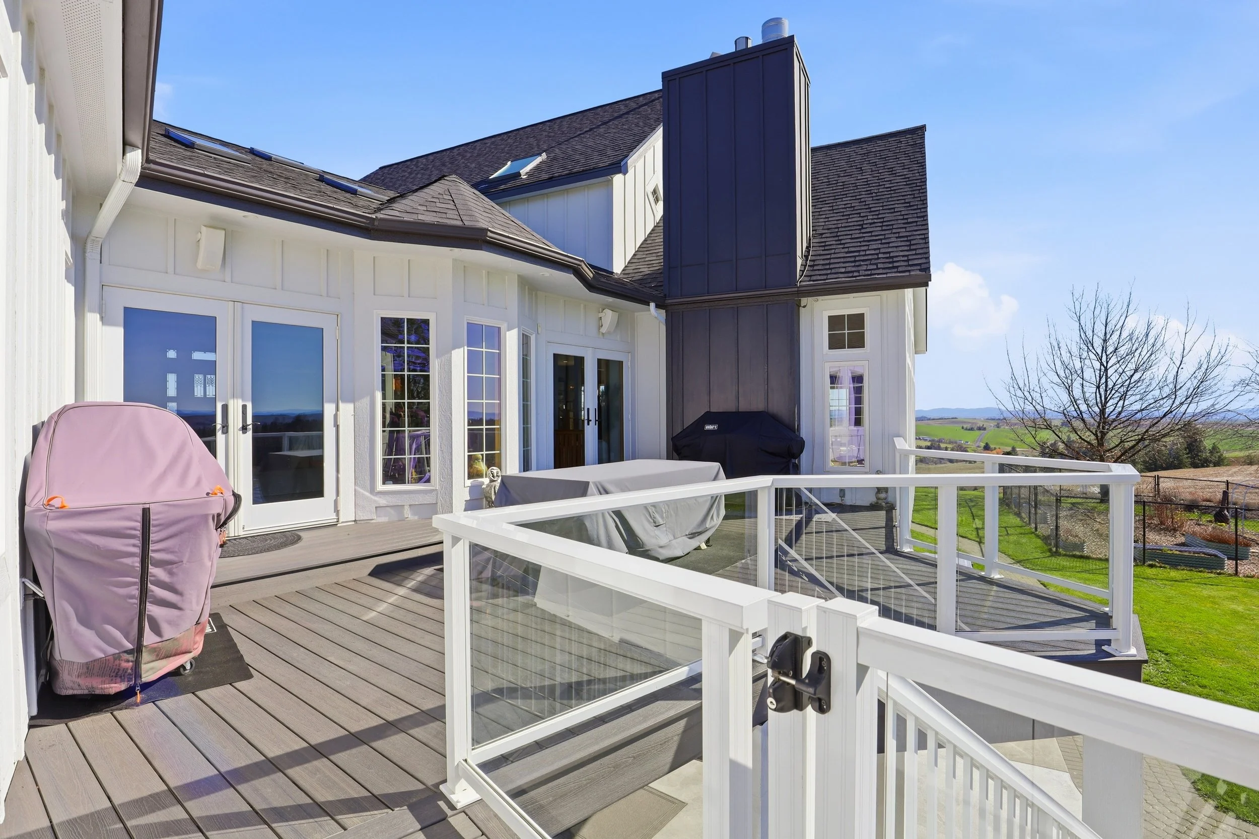 Backyard deck with white railing, a barbecue grill covered with a cover, and a pink backpack on the deck. The house has white walls, glass double doors, and black roofing with solar panels. There is a scenic landscape with green fields and a tree in 