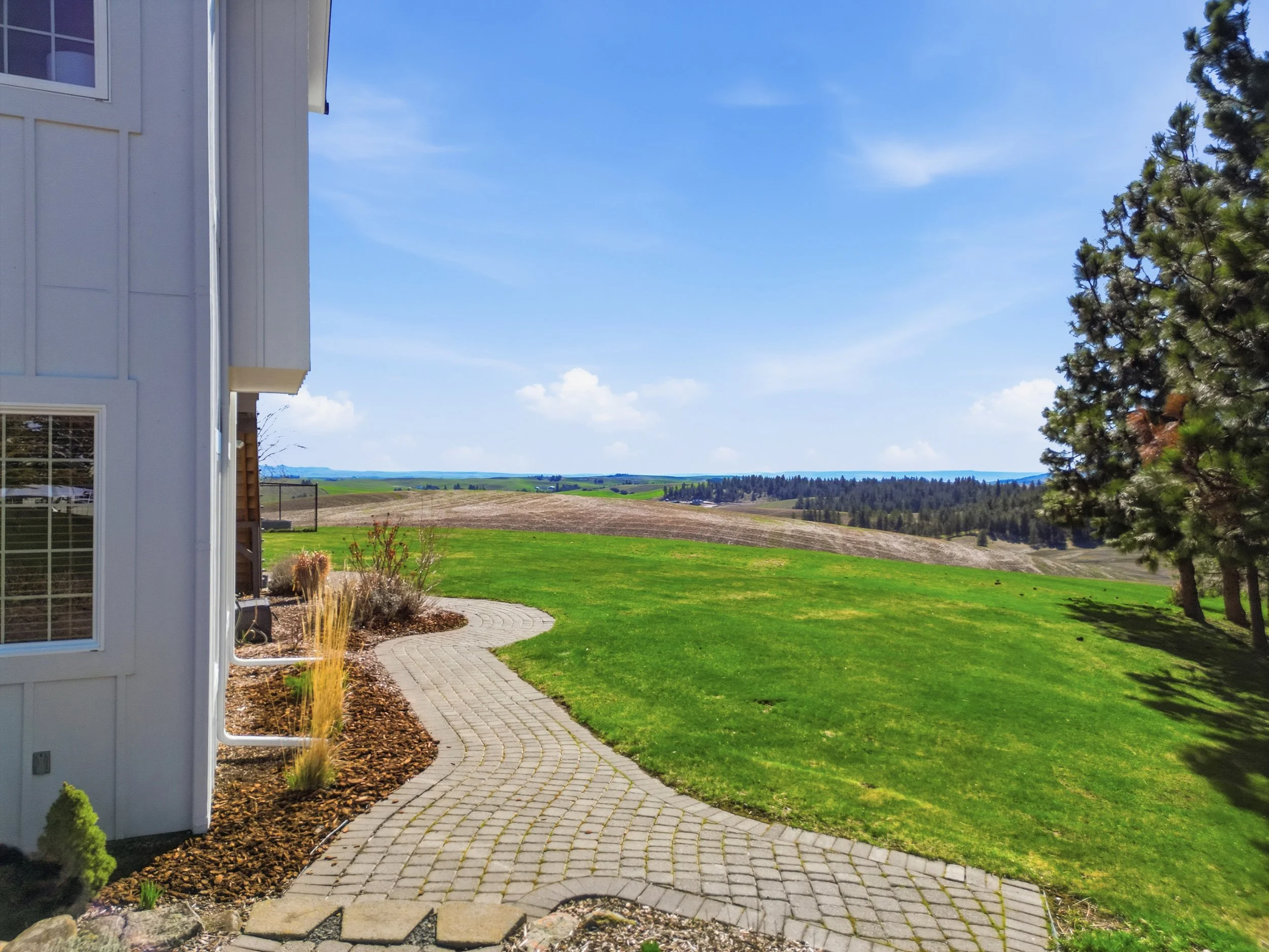 View of a landscaped backyard with a curved paved walkway, a green lawn, and a scenic landscape of rolling hills and blue sky in the background.