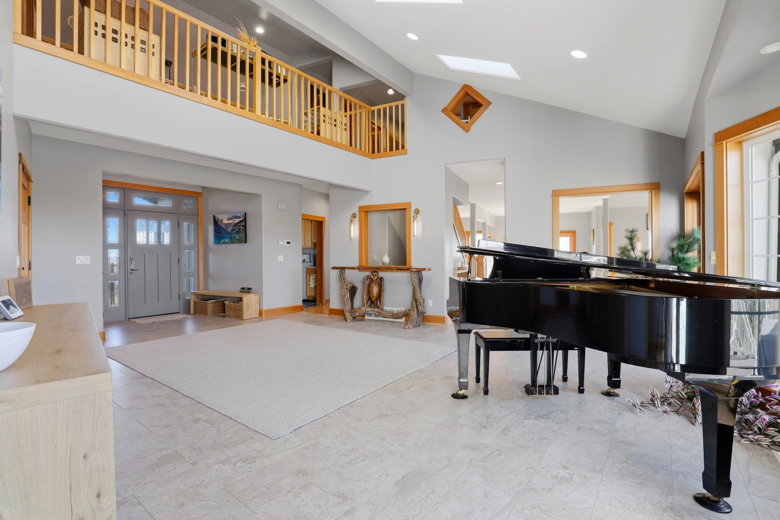 A spacious living room with a grand piano, light-colored tiled flooring, a white and wooden color scheme, and large windows allowing natural light.