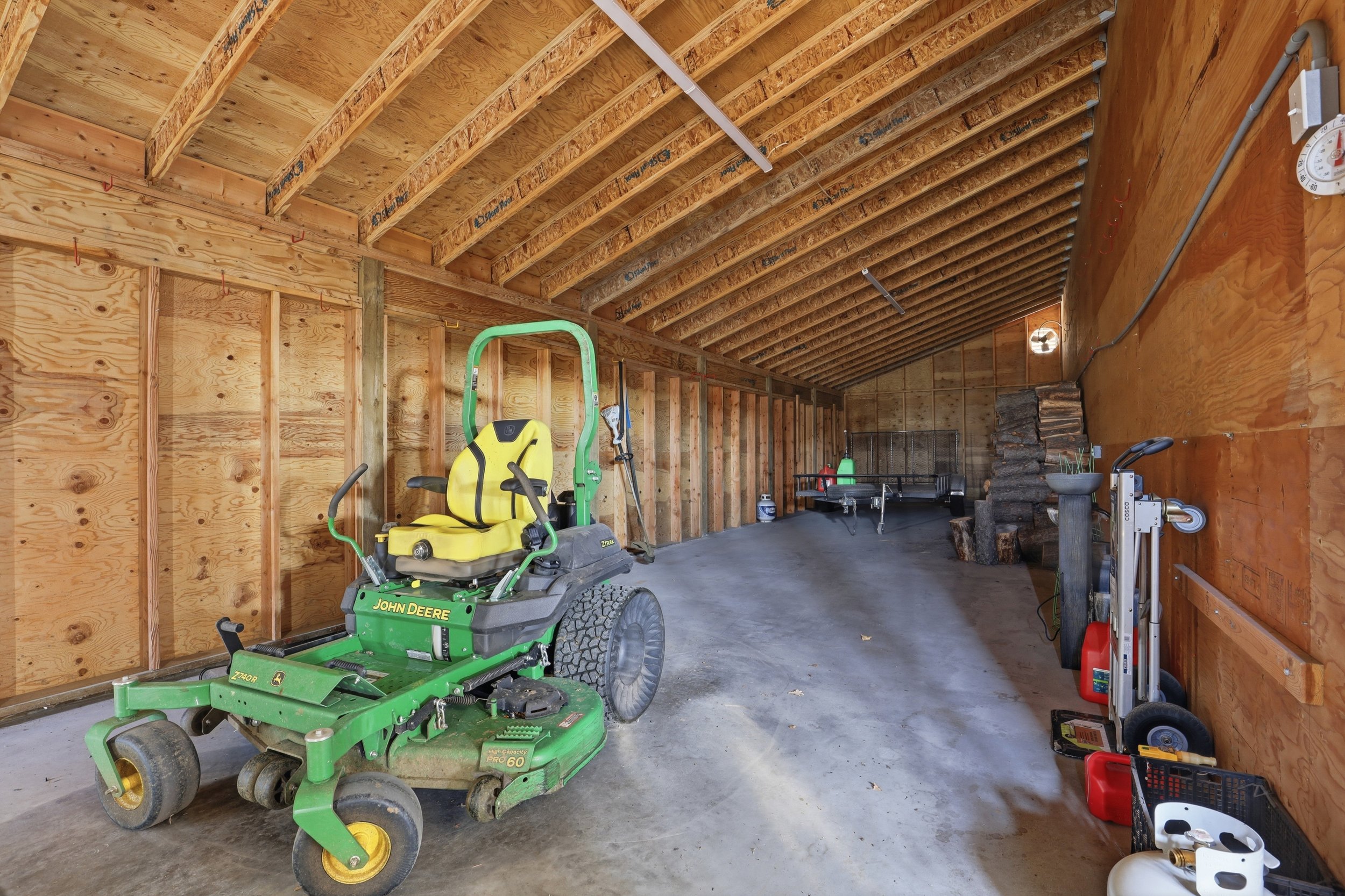 An unfinished garage with wooden framing, a John Deere riding mower, various tools, and firewood stored along the wall.