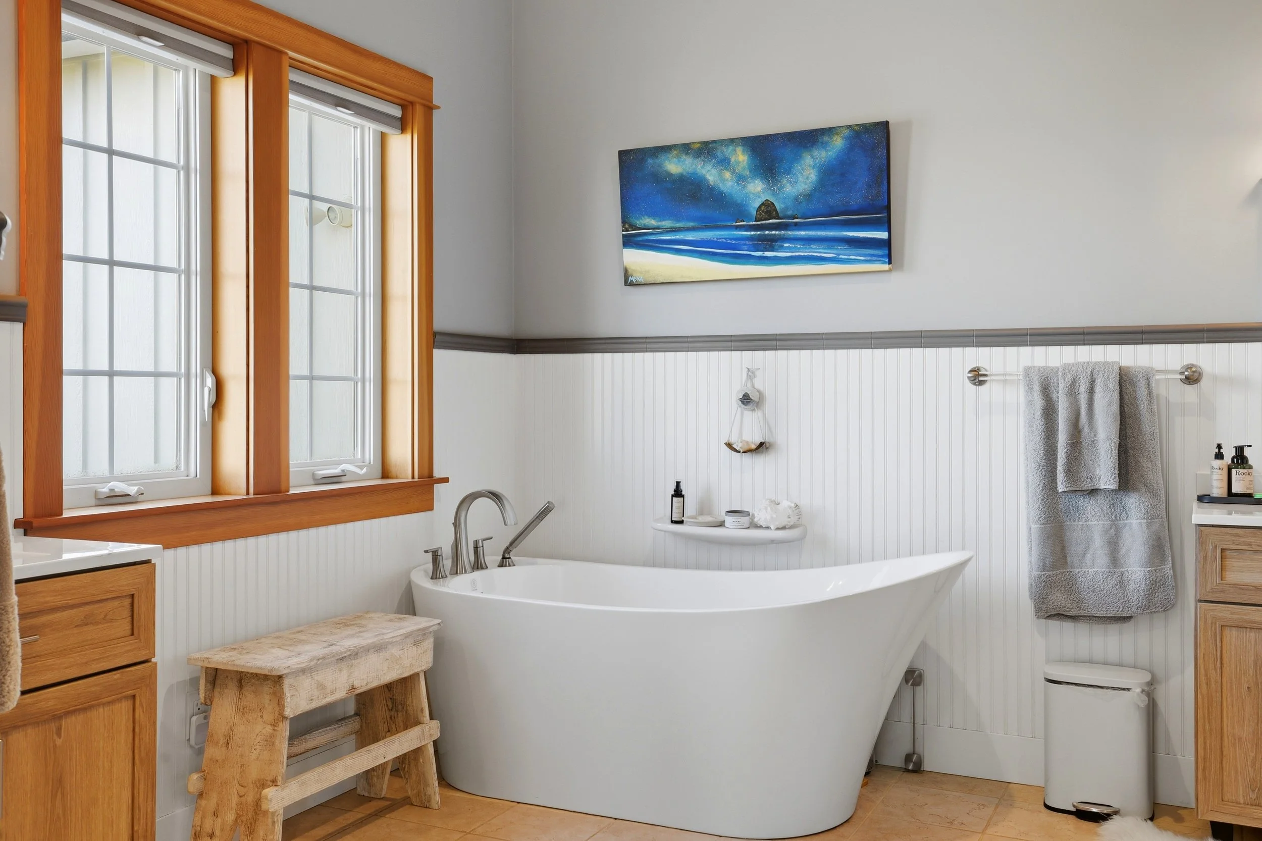 A bathroom with white beadboard walls, wooden trim around two windows, a white bathtub, a wooden stool, a small floating shelf with bath products, a gray towel on a metal towel bar, a painting of a starry night sky over an ocean, and wooden cabinetry