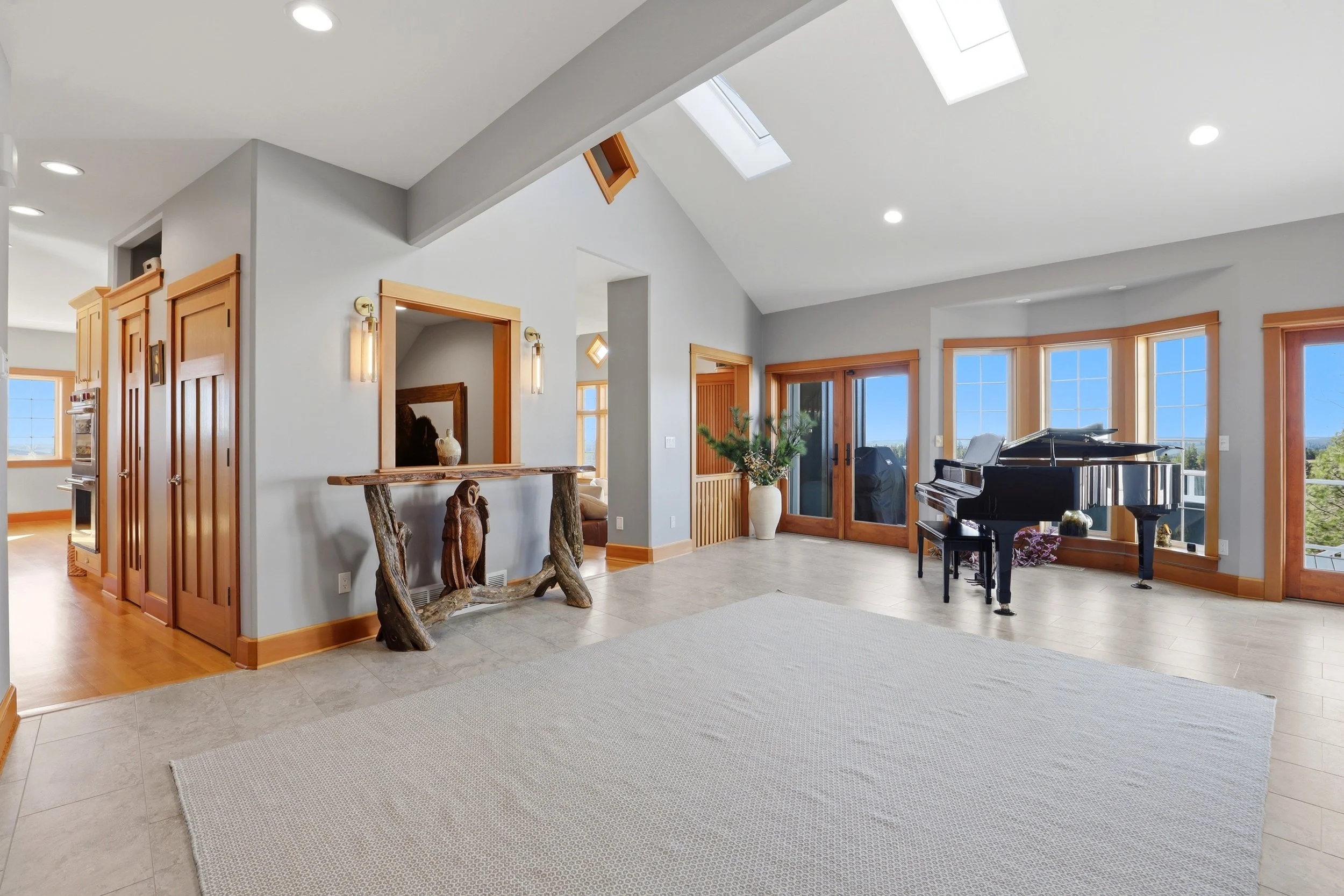 Spacious living room with a grand piano near large windows, a potted plant, a wooden door, and a skylight, with walls painted light gray and hardwood trim.