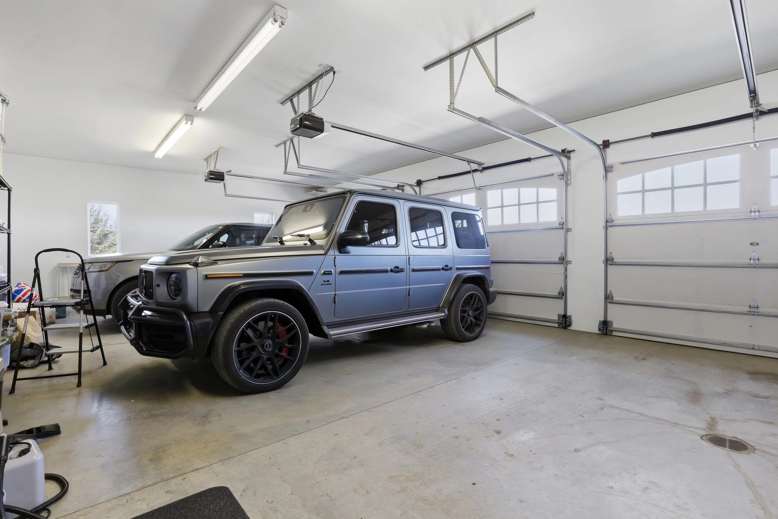 A garage with a silver Mercedes-Benz G-Wagon parked inside, along with another vehicle partially visible. The garage has a concrete floor, a large garage door, and fluorescent ceiling lights. There is some equipment and shelving on the left side.