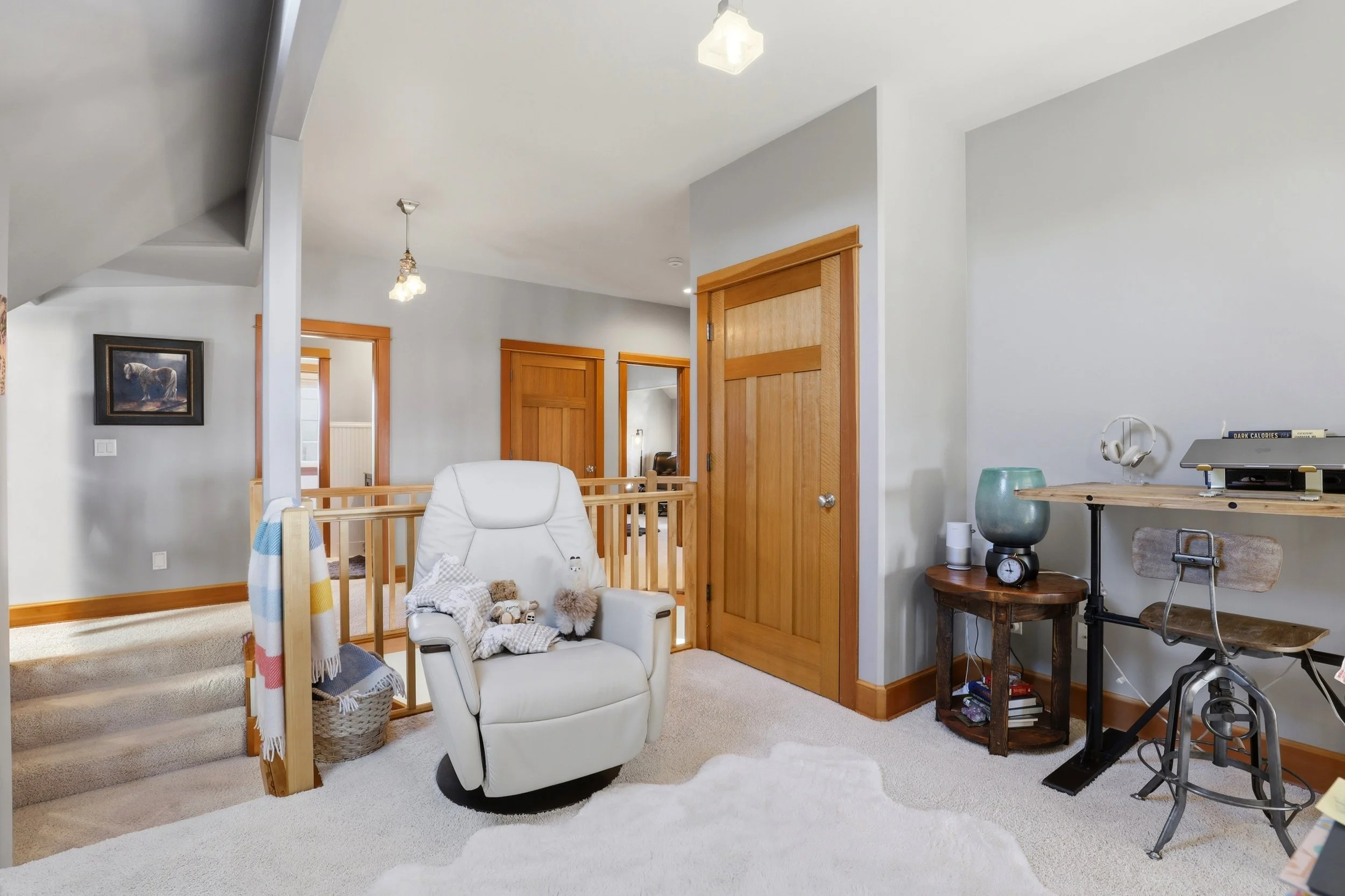 A cozy living room with white walls and beige carpet, featuring a white armchair with stuffed animals, a wooden staircase with a basket at the bottom, and a small workspace with a wooden desk, a clock, and headphones on a stand.
