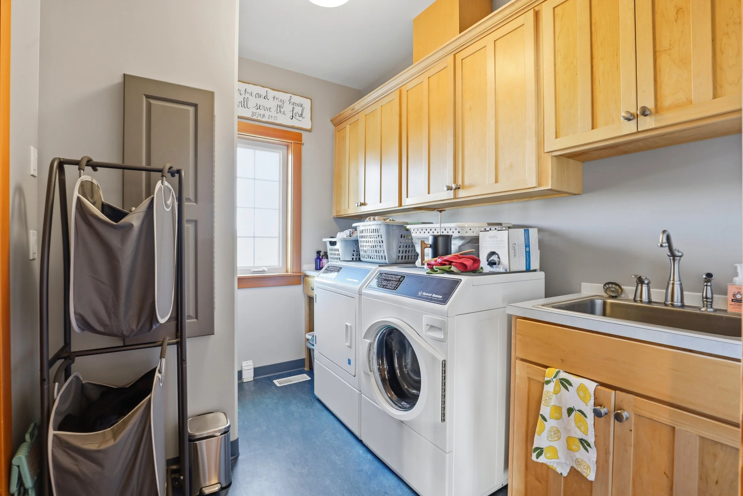 A laundry room with wooden cabinets, a washing machine and dryer, a sink, and laundry supplies. There's a window with a sign above it that reads 'As for me and my house, we will serve the Lord, Joshua 24:15.' Laundry baskets and a drying rack are als