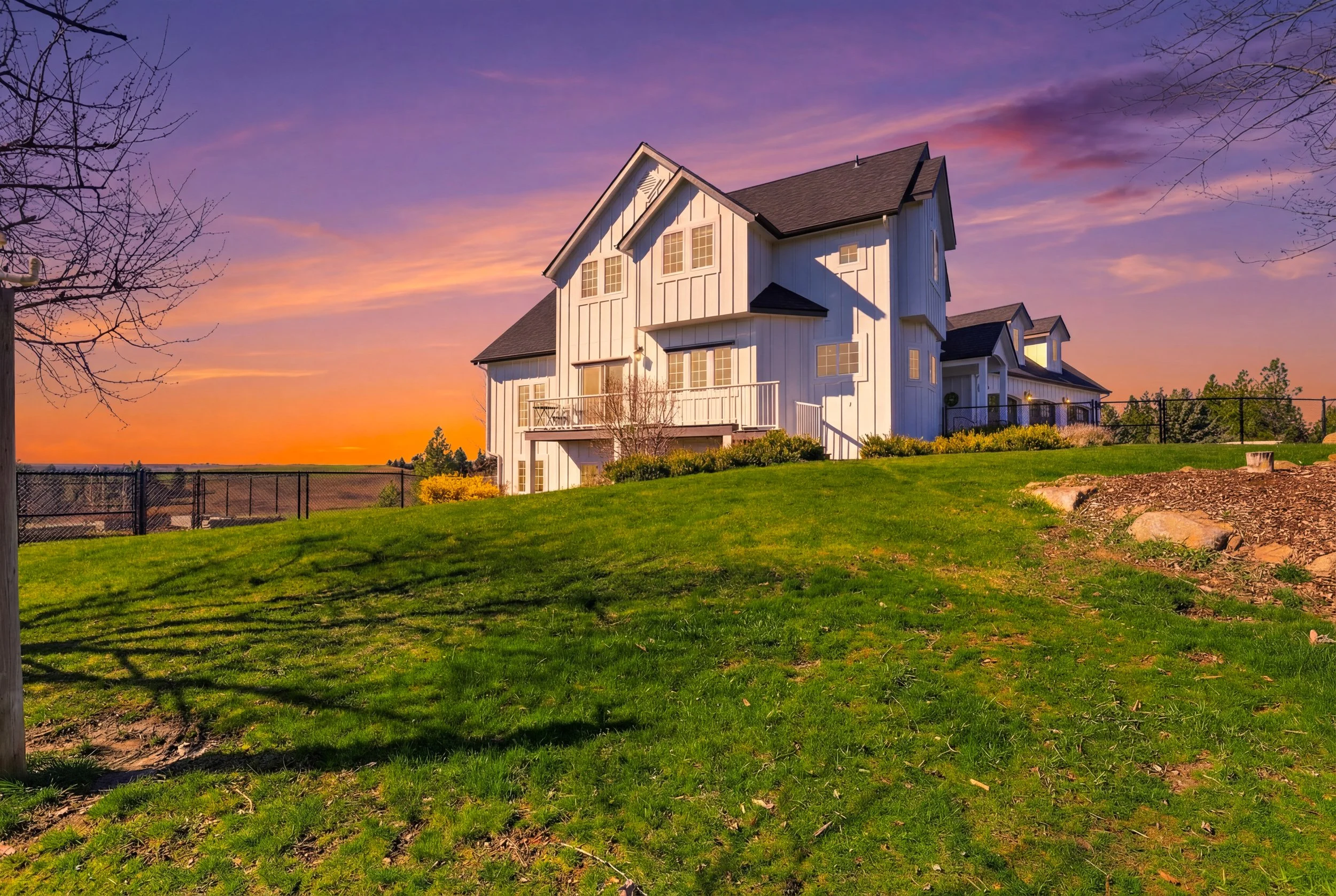 White two-story house with large windows and a balcony, set against a colorful sunset sky with pink and purple clouds, surrounded by a green lawn and some leafless trees.