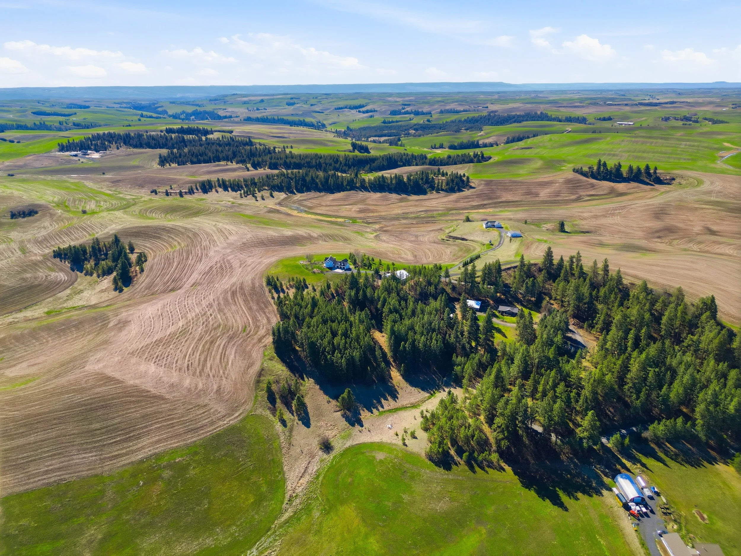 Aerial view of rolling green and brown farmland with scattered trees and farm buildings on a sunny day.