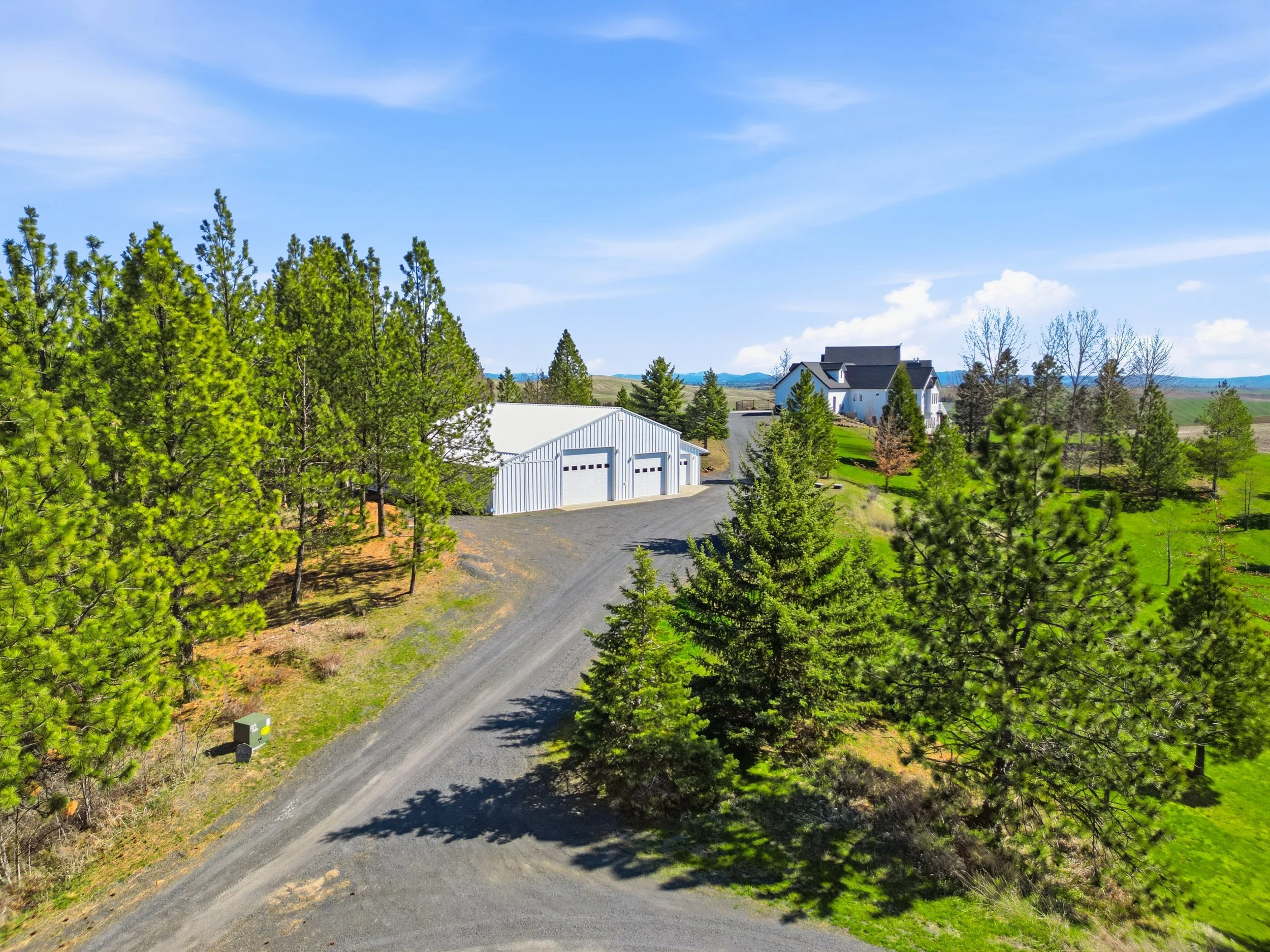 A rural landscape with a gravel road, green trees, a white metal building, and houses in the distance under a blue sky with scattered clouds.