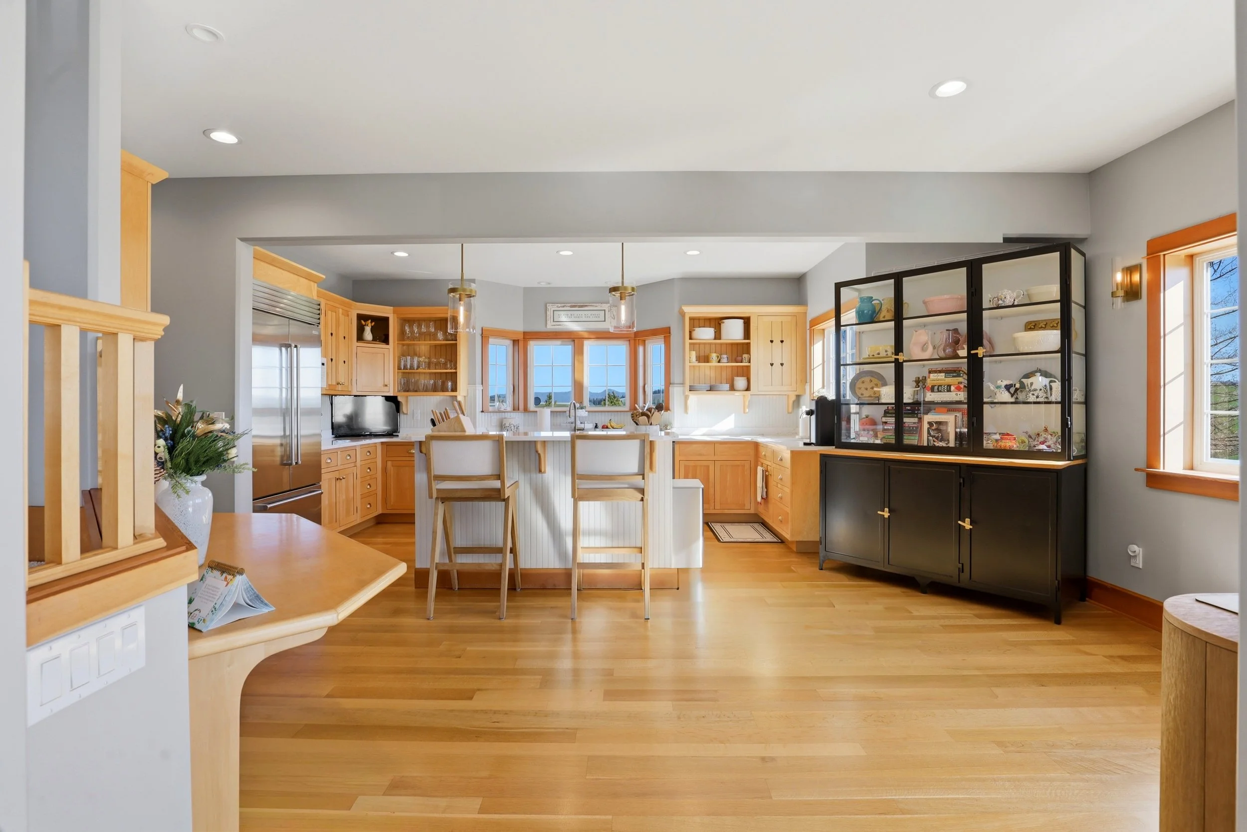 Open-concept kitchen with wooden cabinetry, a central island with barstools, and large windows showing a view outside.