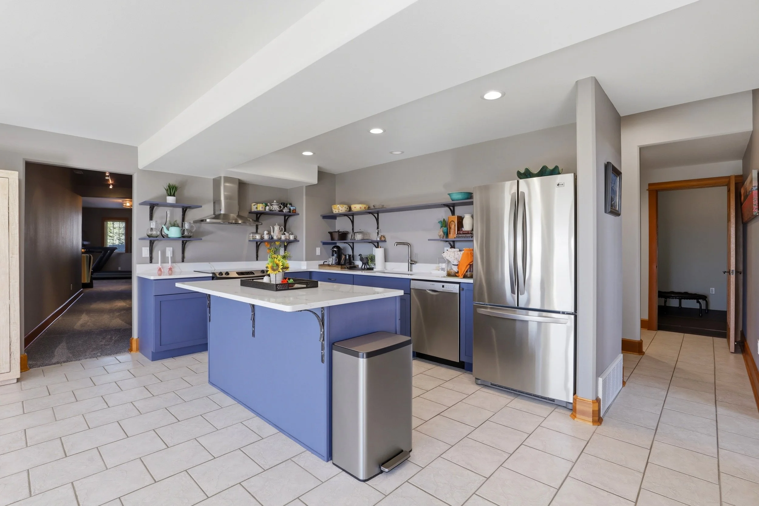 Modern kitchen with blue cabinets, stainless steel appliances, open shelving, island, tiled floor, and recessed lighting.