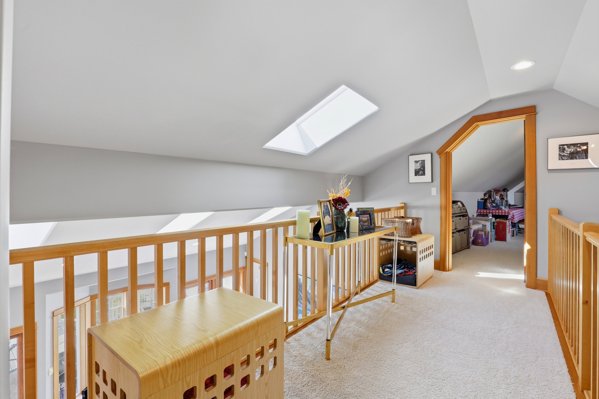 An interior view of a two-story home with a sloped ceiling and a large skylight window. The area is decorated with framed photos, a flower arrangement, and candles on a table. A wooden railing separates this upper level from the lower level, which is