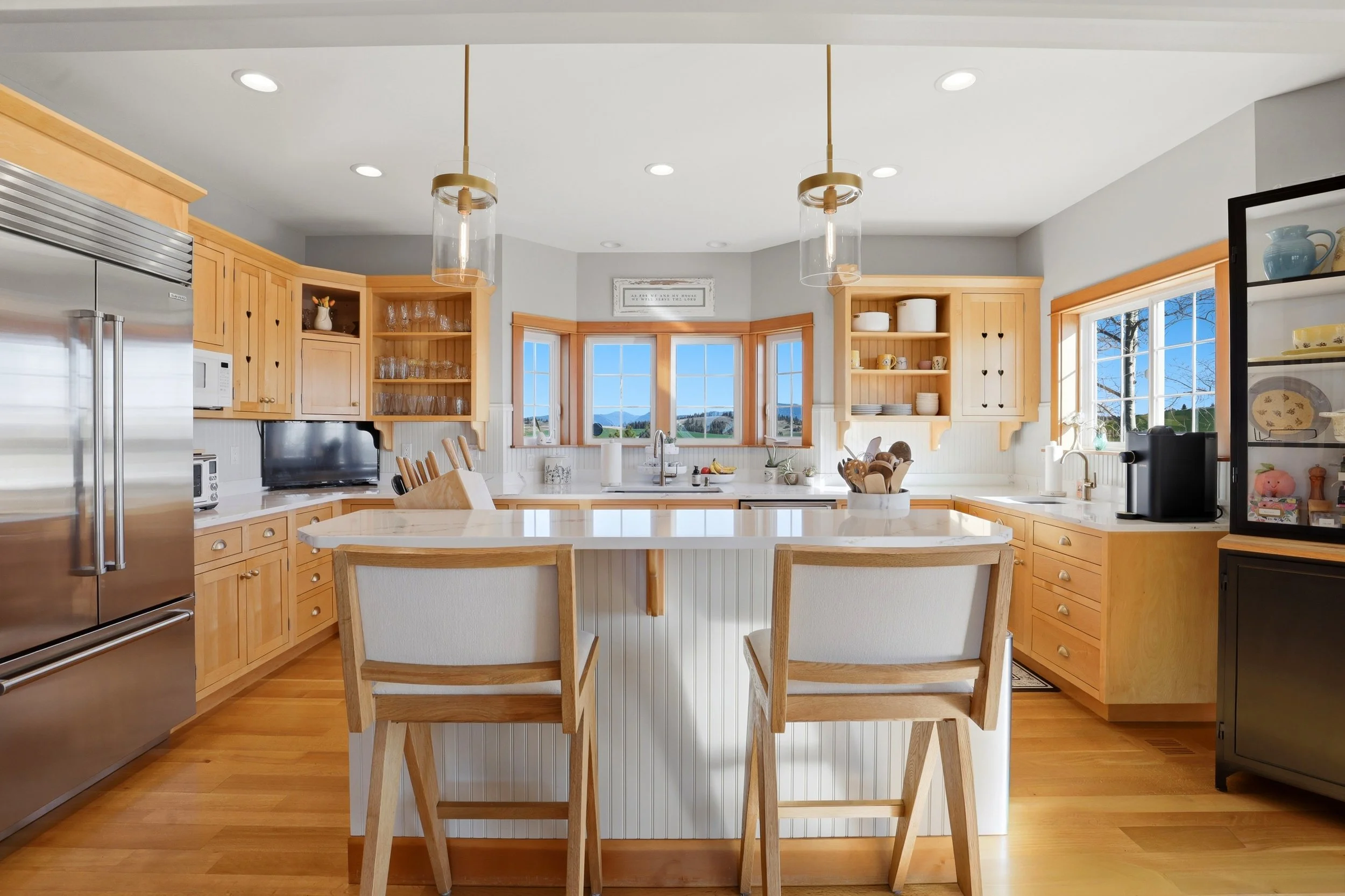 A bright kitchen with large windows showing a scenic outdoor view, featuring natural wood cabinets, a white kitchen island, and modern appliances.