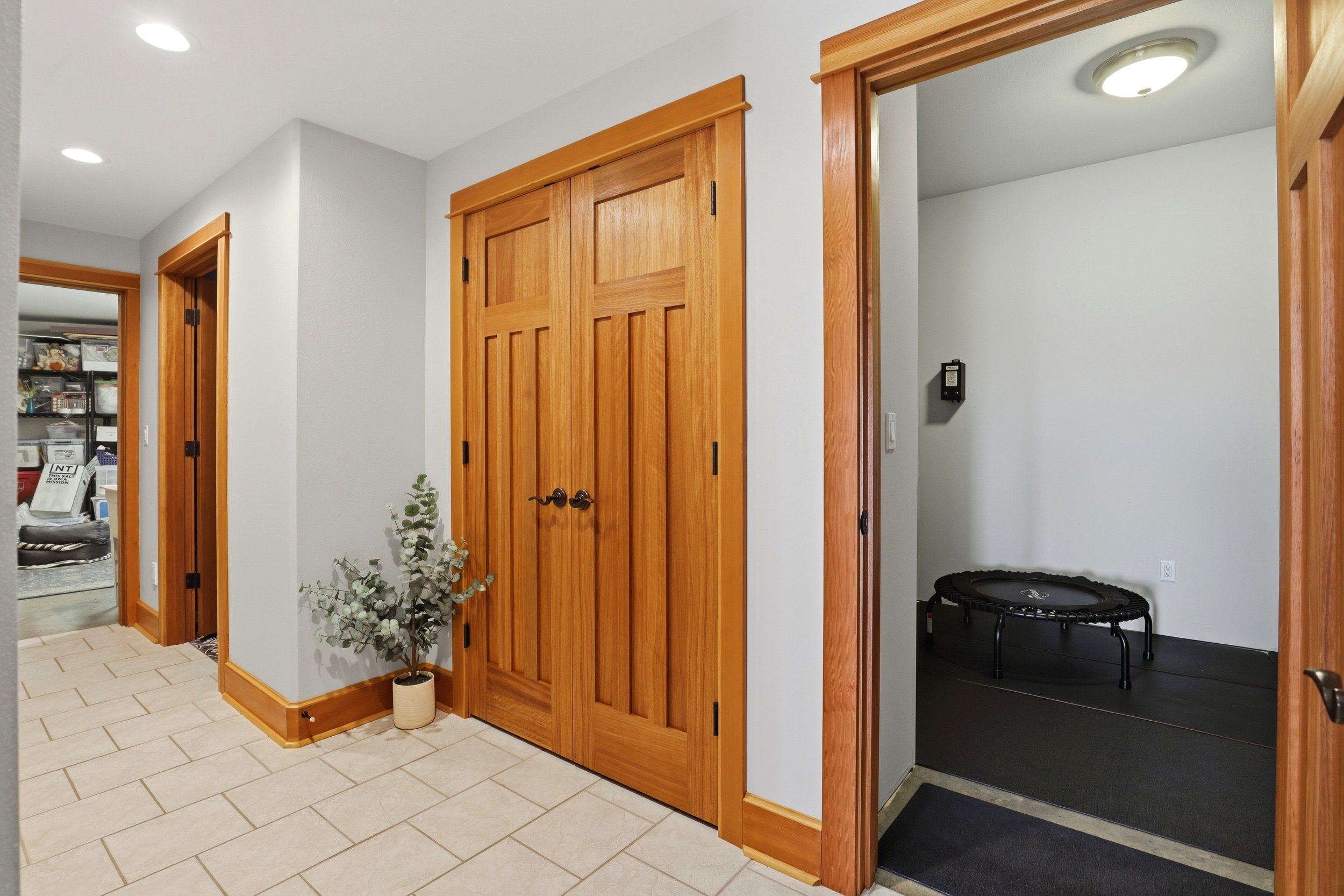 Interior view of a hallway with wooden doors, tiled floor, a potted plant, and a small trampoline in a room.