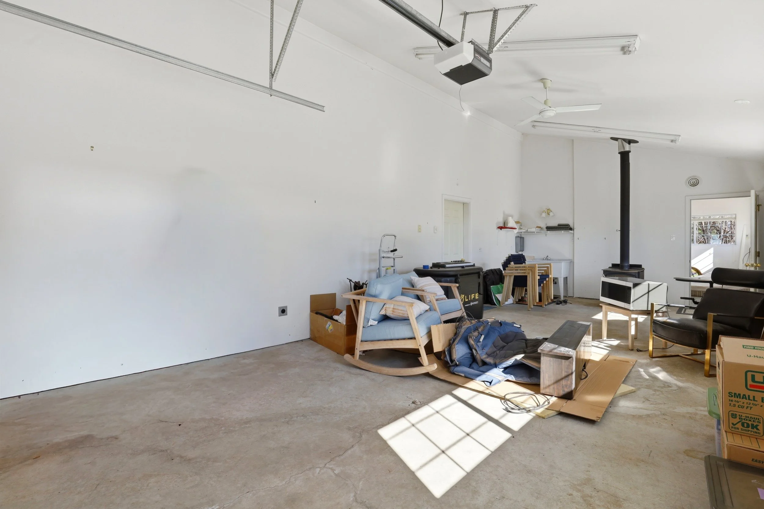 Living room with clutter, including chairs, boxes, and a couch, illuminated by sunlight through a window, with a white wall and garage ceiling.