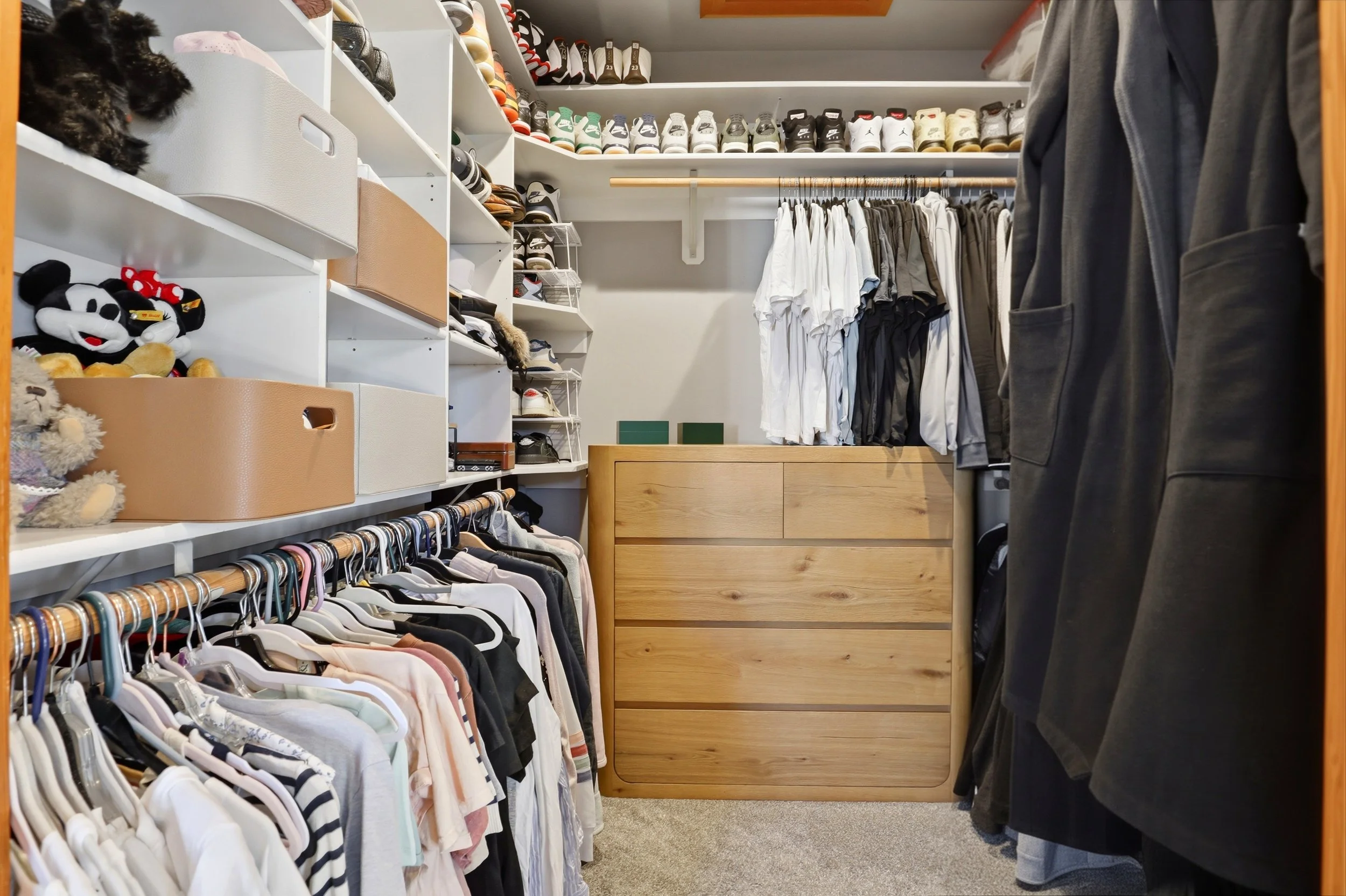 A walk-in closet with shelves filled with sneakers on the top, hats, and clothing on hangers, with a chest of drawers at the back and a person wearing dark clothes standing to the right.