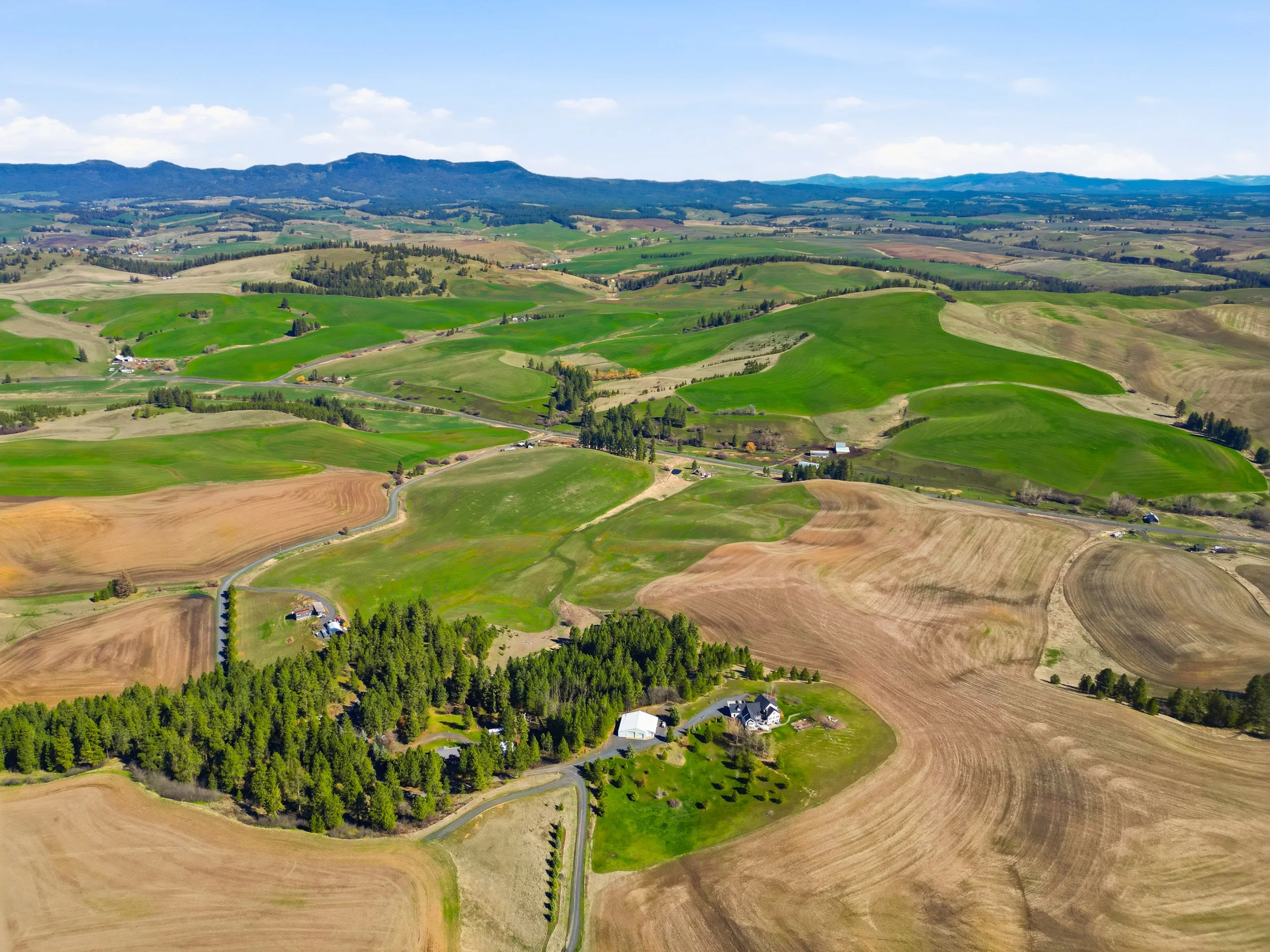 Aerial view of rolling green hills, farmland, forests, and scattered houses under a bright blue sky with some clouds.