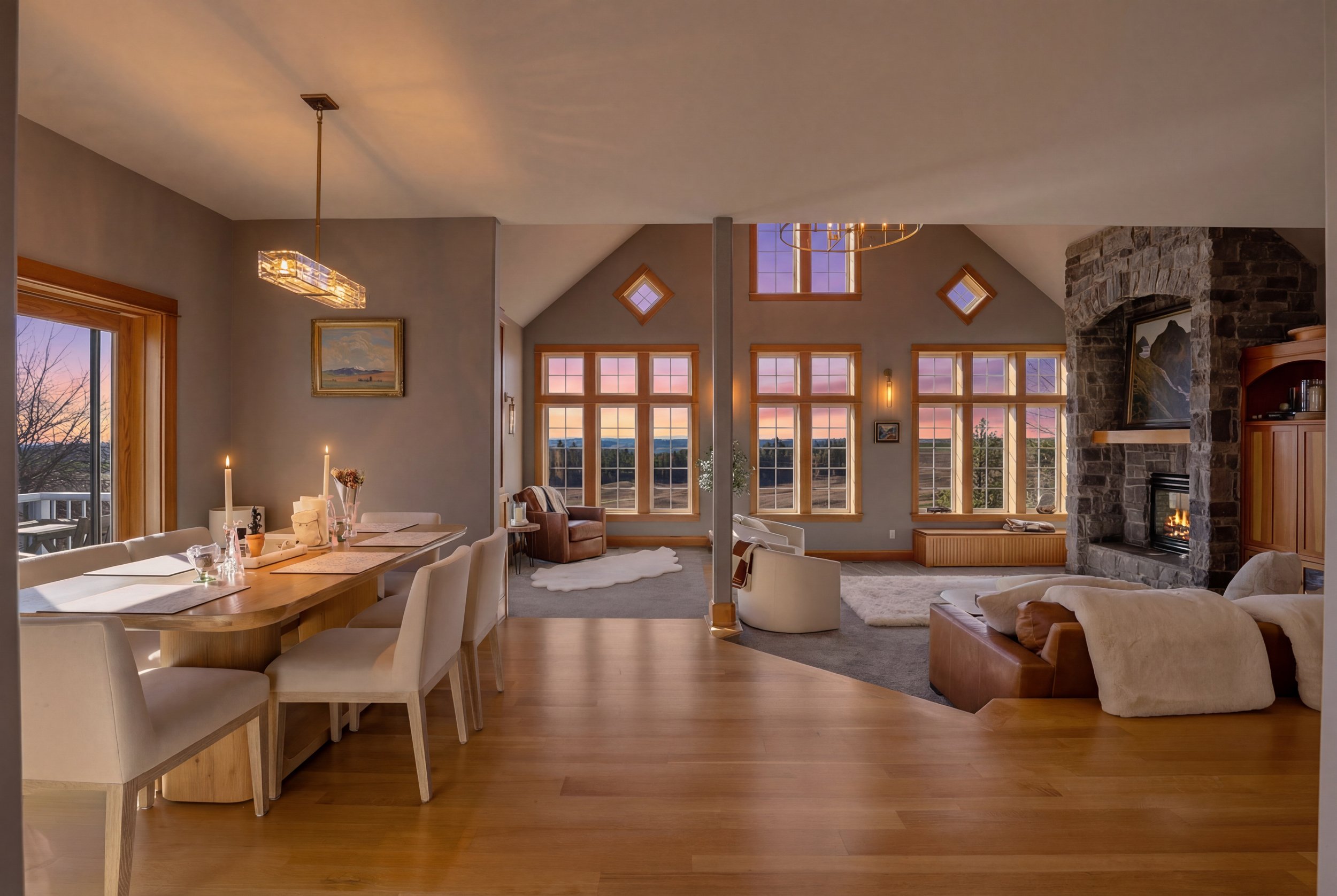 Spacious living and dining area with large windows showing sunset view, stone fireplace, modern chandelier, and cozy furniture.