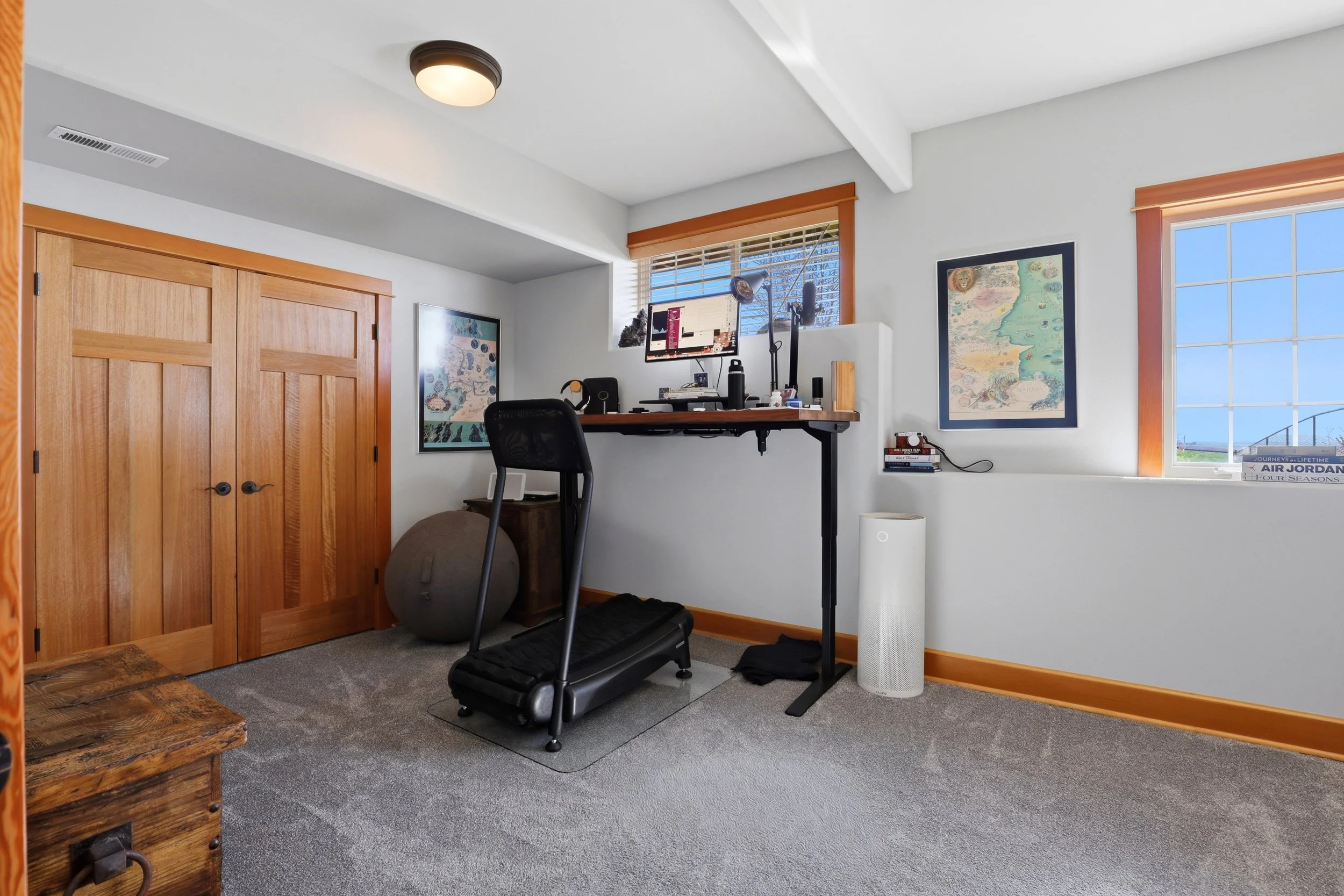 Home office room with white walls, wooden door and window trim, a window with blinds, a wall-mounted desk with computer monitor, microphone, and speaker, a black chair, artwork on the walls, a small stack of books, a white air purifier, and a treadmi