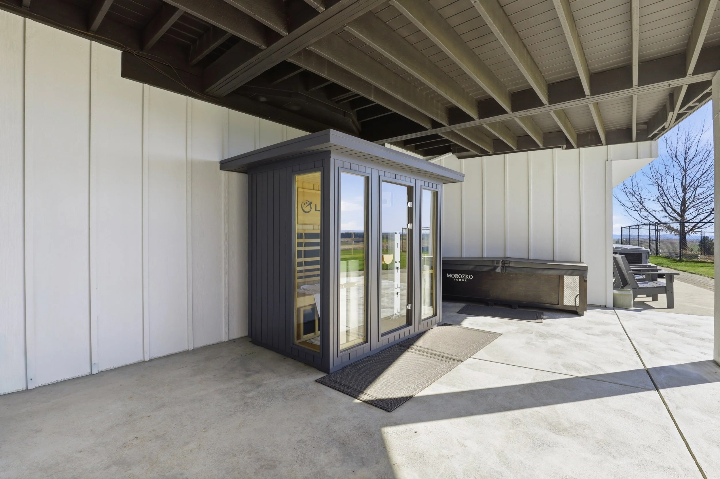 Outdoor patio area with a modern glass-enclosed sauna, a large grill or smoker, and a patio bench. The patio is covered by a large wooden overhang, with white siding walls and a concrete floor. In the background, there is a fenced backyard with a tre