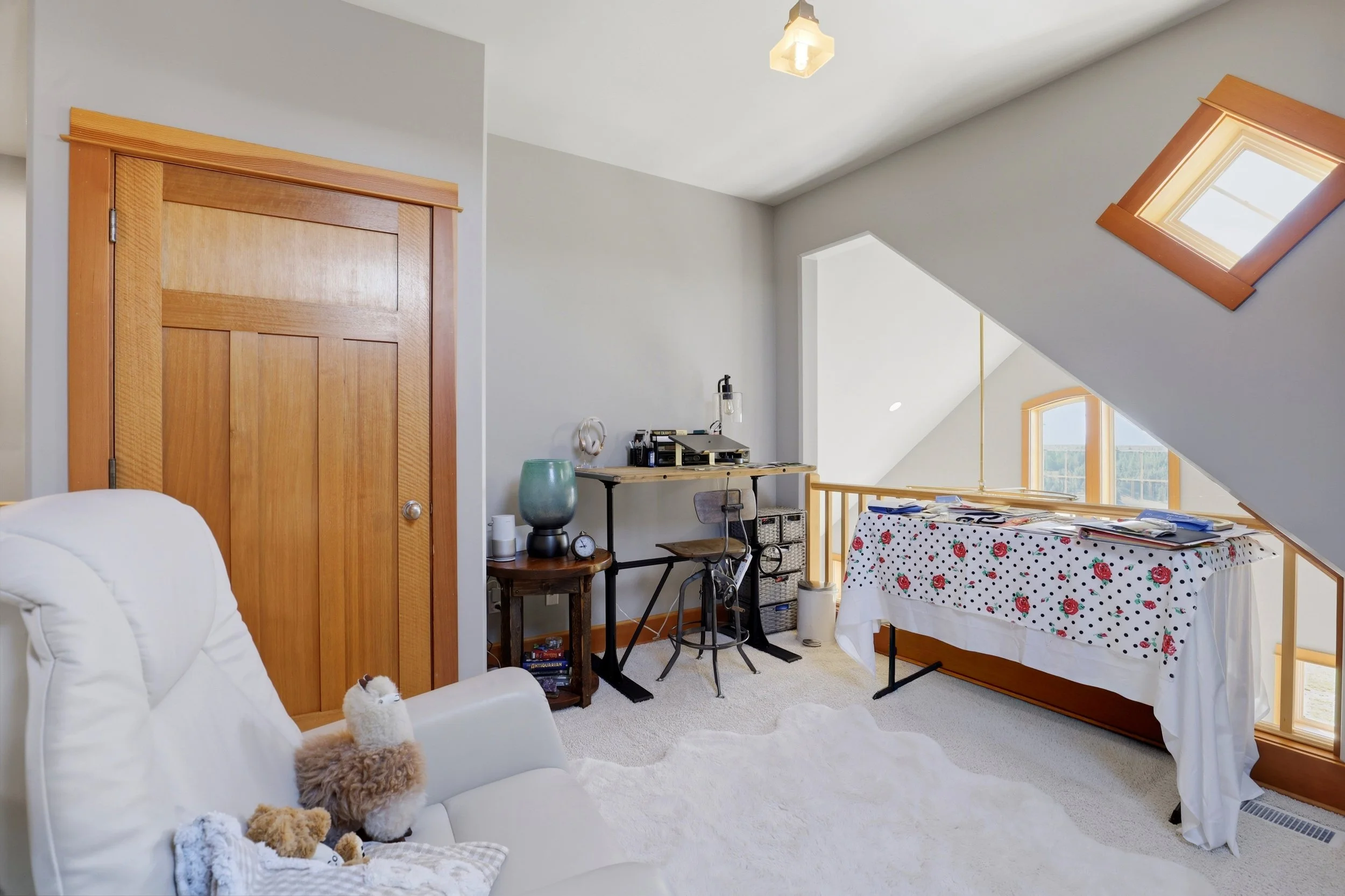 A cozy attic bedroom with a sloped ceiling and skylights. The room features a white armchair with stuffed animals, a green vase, a small wooden table with a clock, and a desk with office supplies. A bed with a floral bedspread is partially visible.