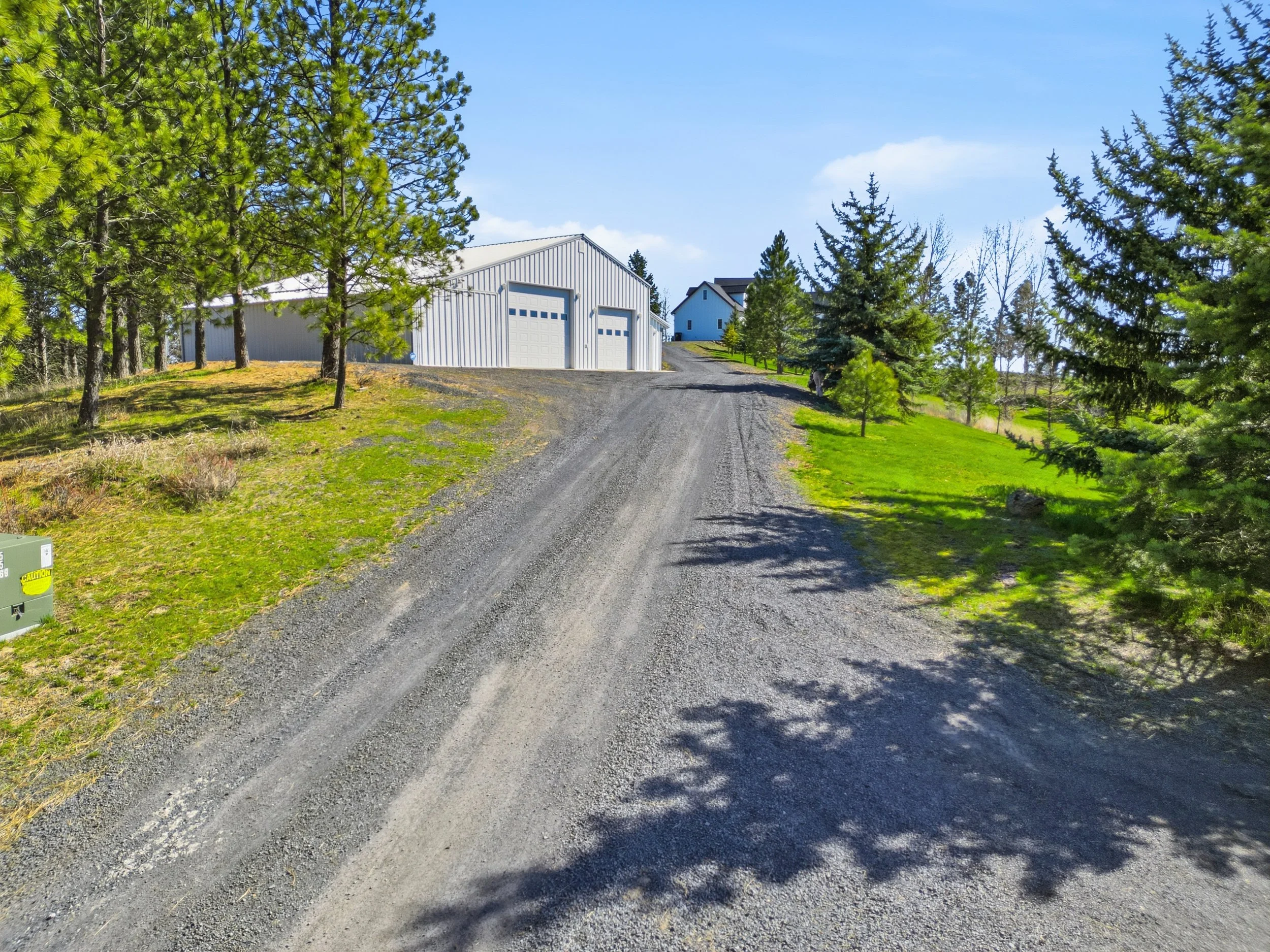 A gravel driveway leading up a slight hill with a large white garage building at the top, surrounded by green grass and tall pine trees under a blue sky with some clouds.