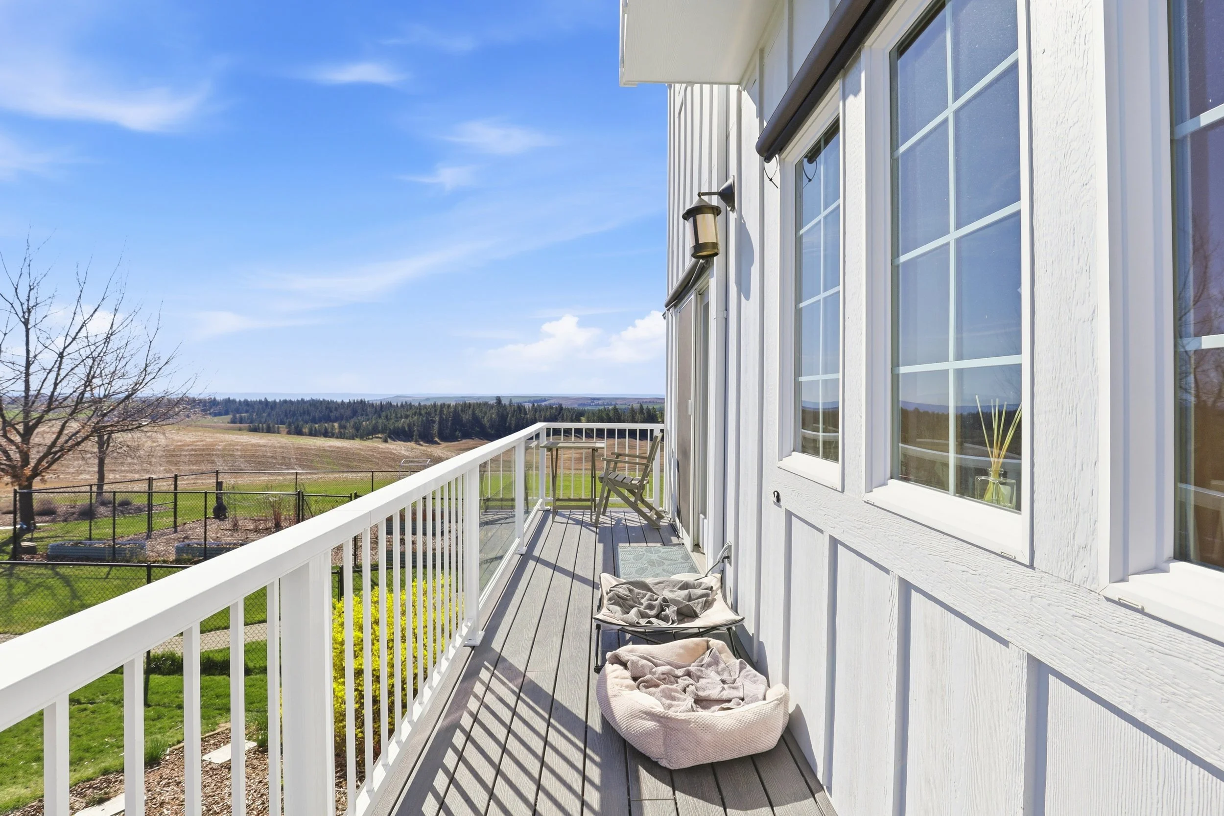 View of a white house balcony with gray wooden flooring, overlooking a rural landscape with fields, trees, and distant hills on a clear sunny day. There is a chair and some blankets on the balcony.