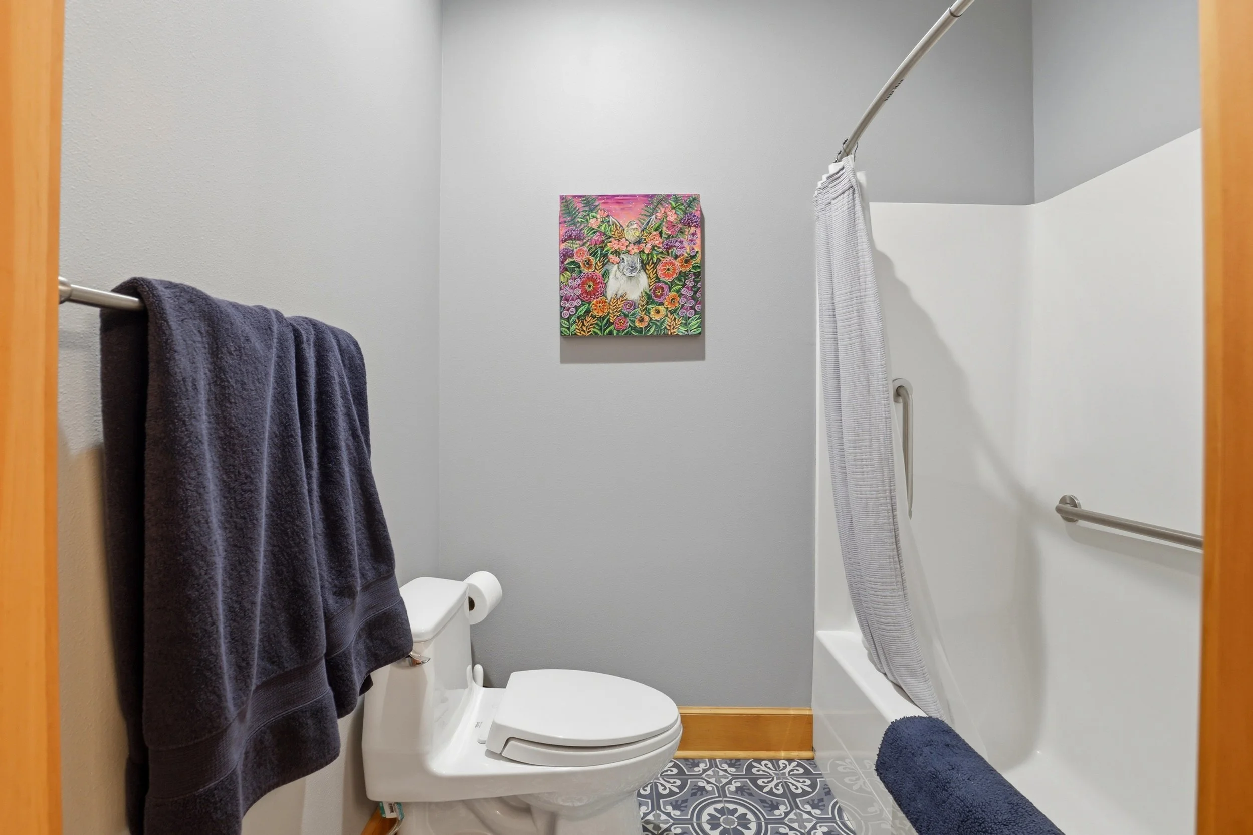 A small bathroom with a white toilet, dark purple towels on a towel rack, a colorful floral art piece on the wall, a gray shower curtain, and a grab bar on the wall next to the bathtub.