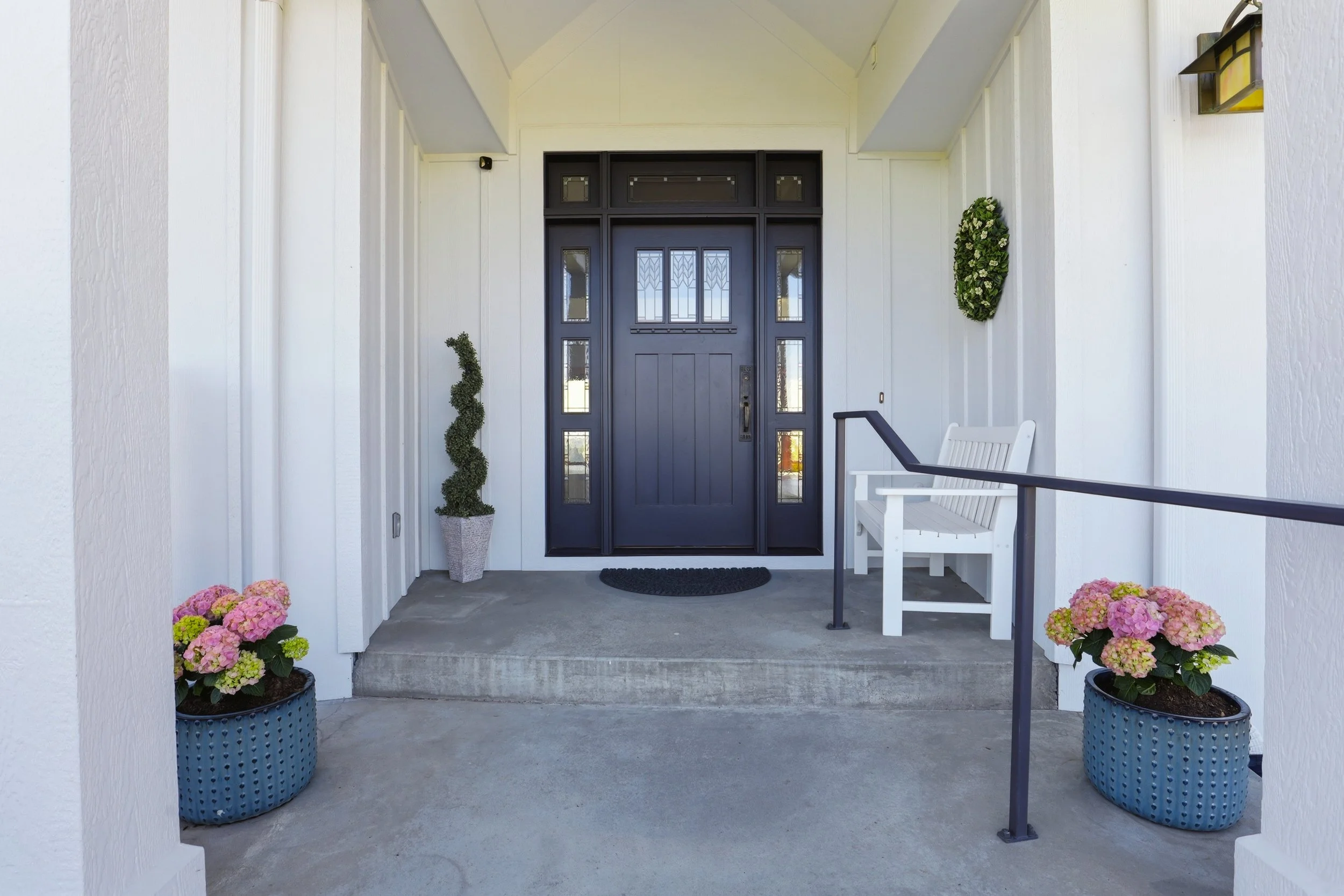 Front porch with a black front door, two potted pink hydrangeas, a white bench, and a wreath on the wall.