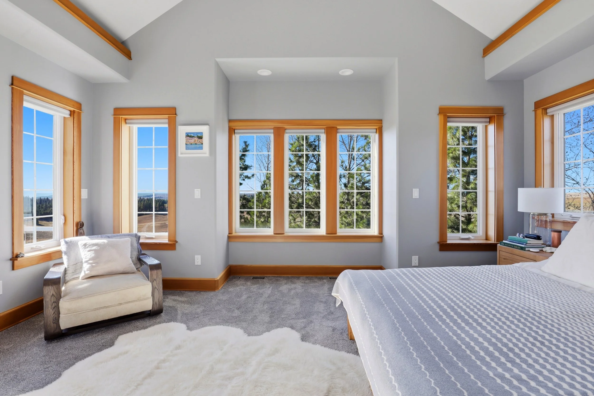 Interior view of a bright bedroom with large windows showing trees and a view outside. There is a cozy armchair with a pillow, a white fluffy rug, and a bed with a striped bedspread. The room has light gray walls, wooden window frames, and trim, with