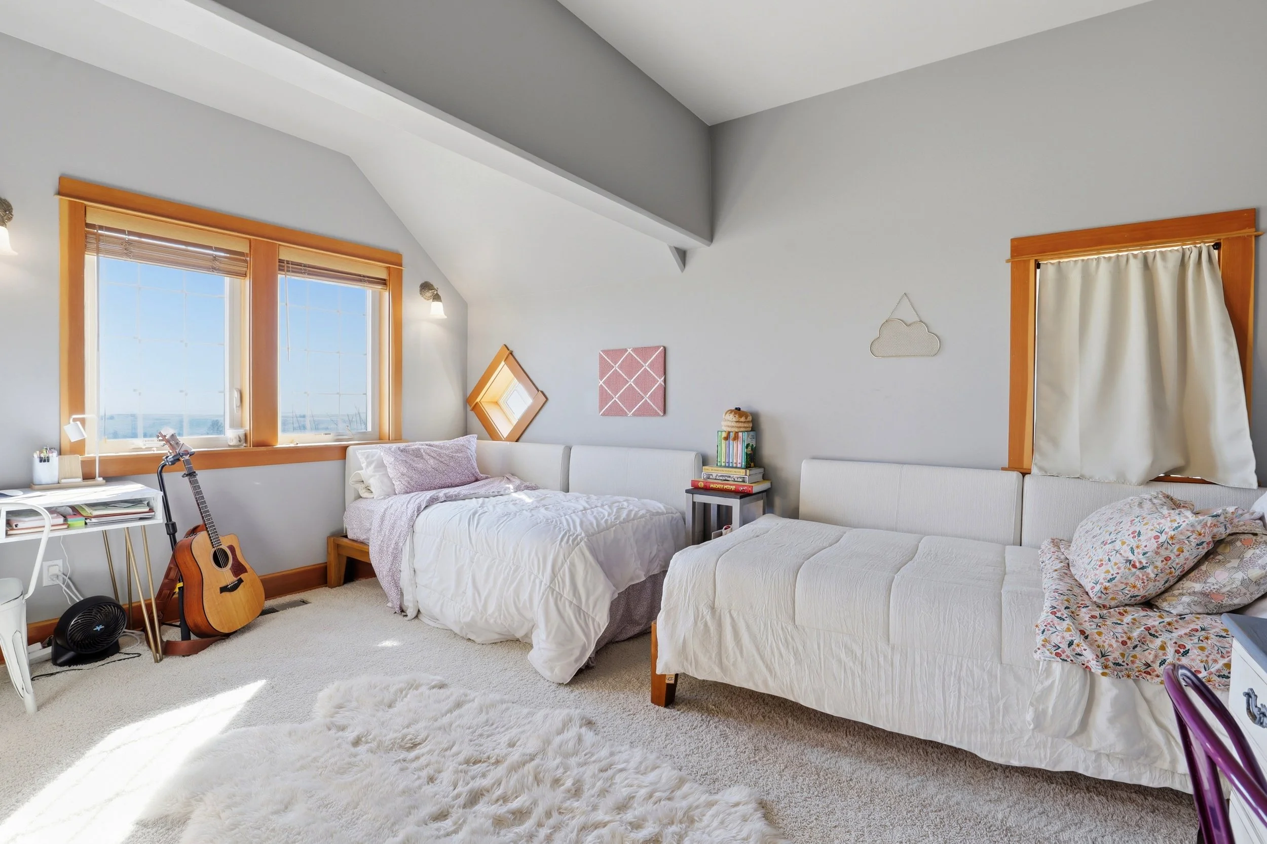 A cozy bedroom with two beds, large windows with wooden frames, a guitar on a stand, a small white table, hanging decor on the wall, and a fluffy white rug on the carpeted floor.