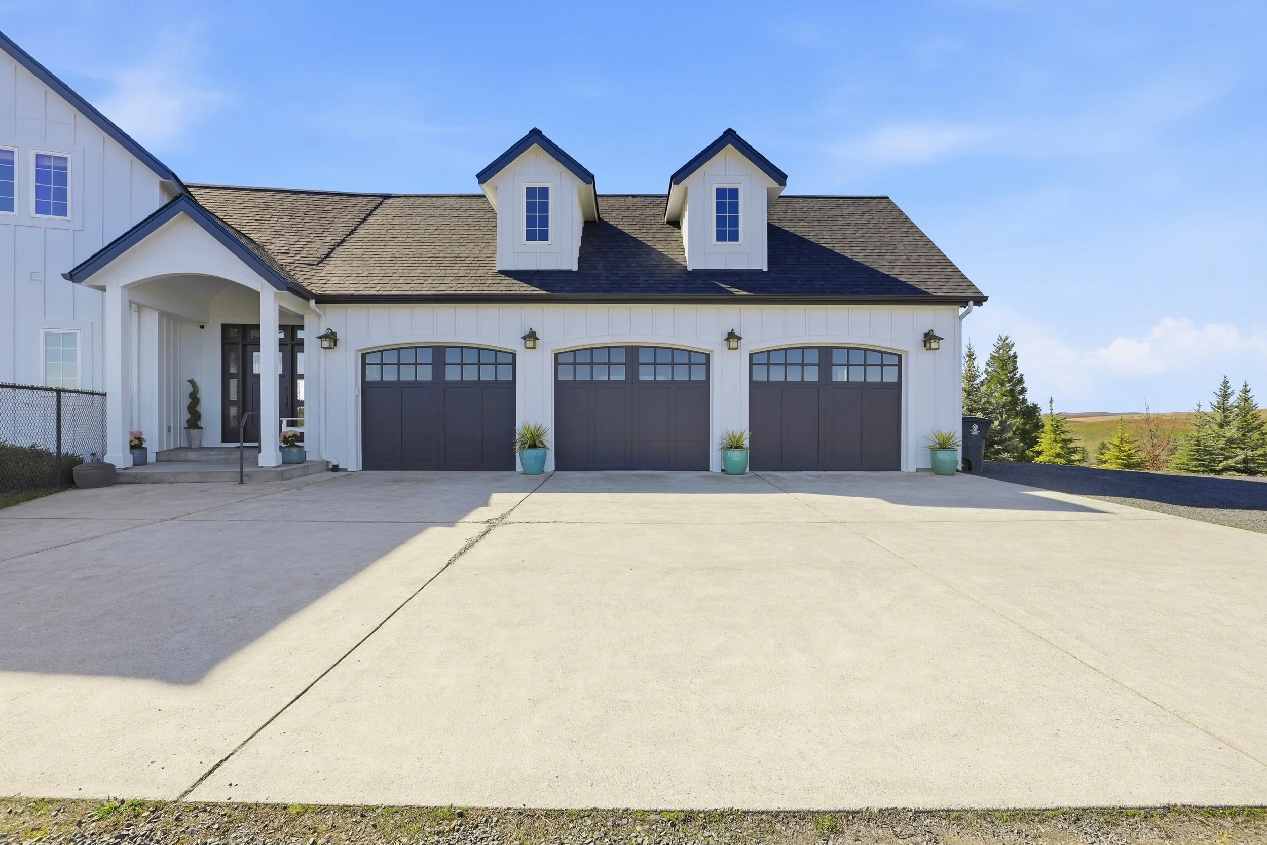 Front view of a modern white house with a three-car garage, two dormer windows, and decorative potted plants on the driveway.
