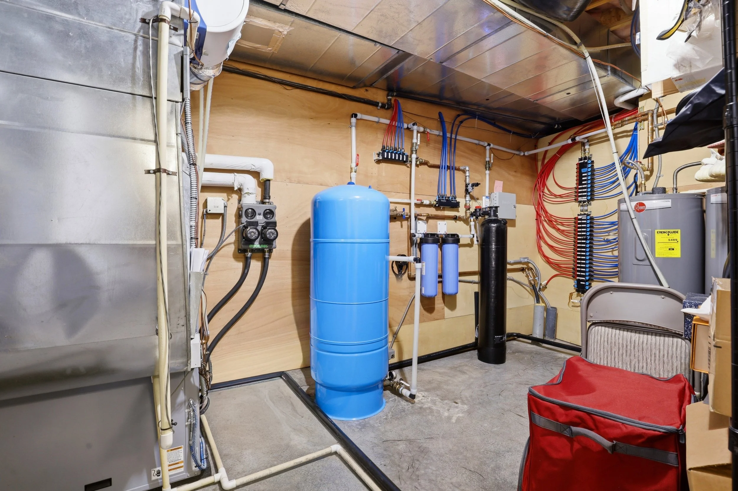 A utility room with various plumbing and electrical equipment, including a large blue water filter tank, pipes, valves, electrical panels, and a water heater, with a wooden wall background and a concrete floor.