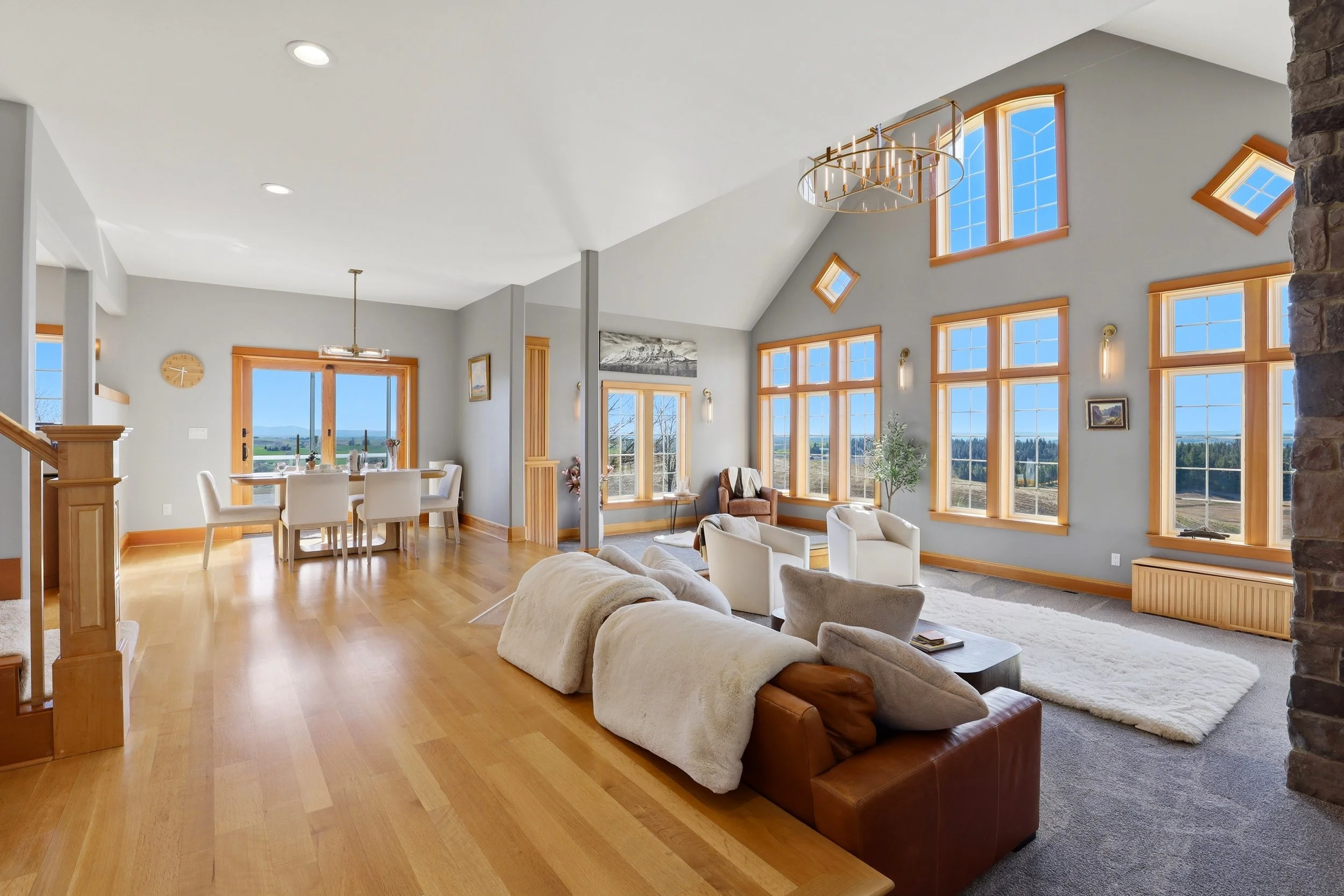 Open living room with large windows, white sofas, a stone accent wall, and a view of the outdoors, featuring a wooden dining table and a chandelier.