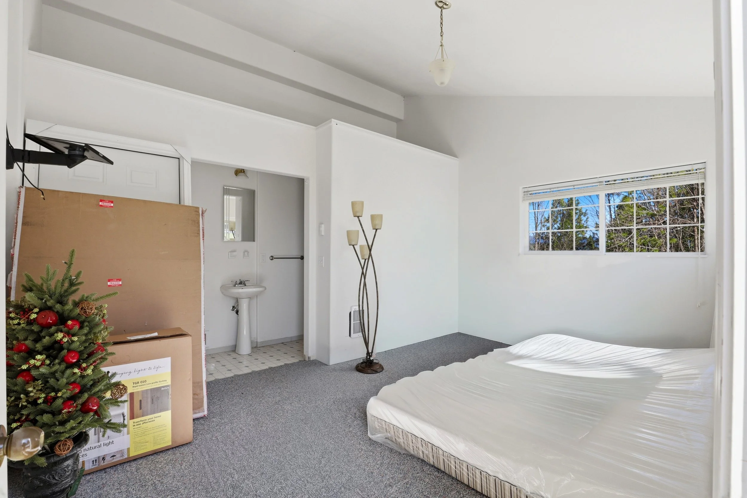 Empty bedroom with a window showing trees outside, a corner with a lamp, a mattress in plastic, a small sink, and packed Christmas tree decorations.