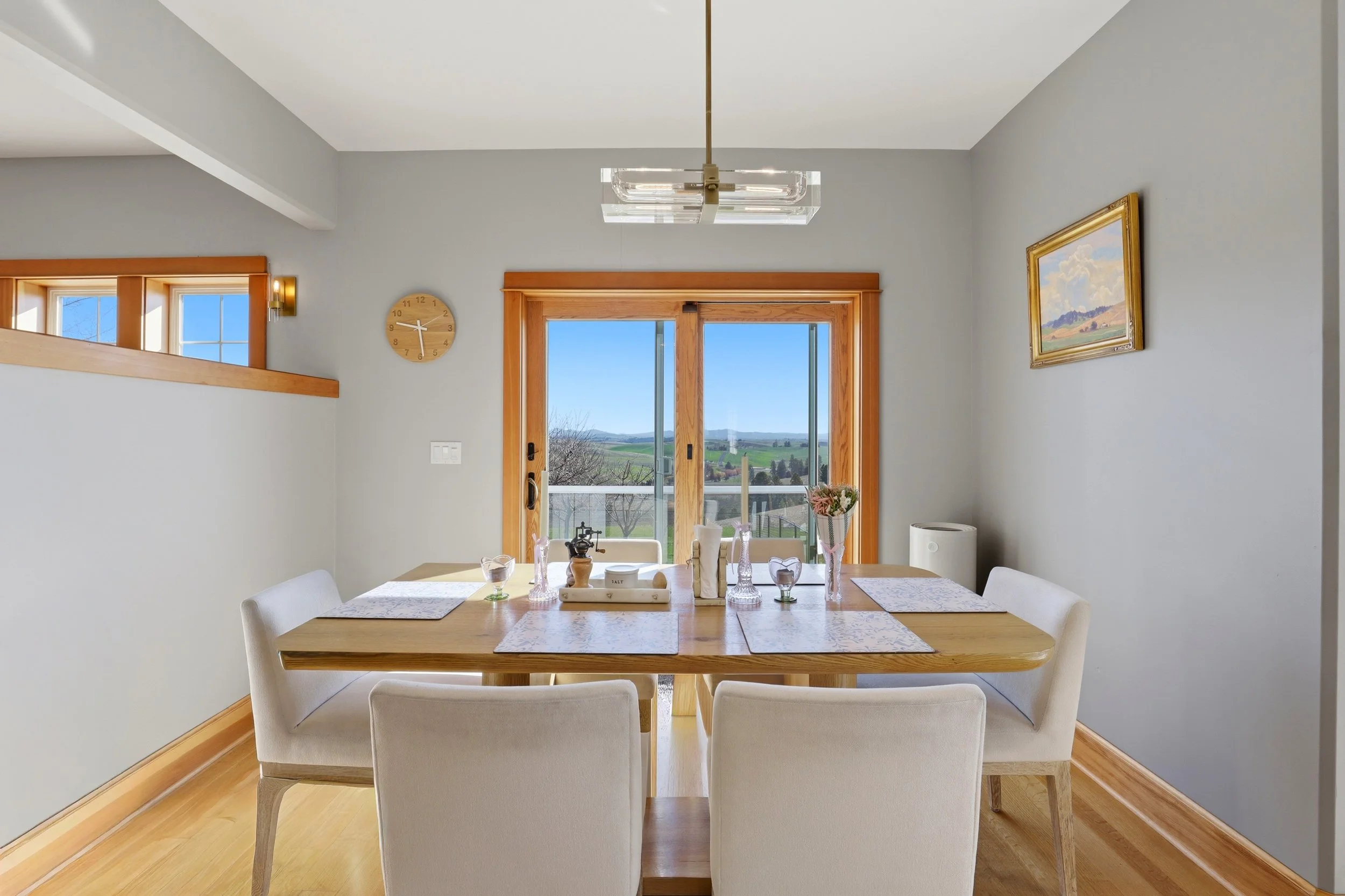 A dining area with a wooden table surrounded by white chairs, placed in front of a large window with a scenic outdoor view. The scene includes a wall clock, a piece of artwork, and a modern chandelier hanging from the ceiling.