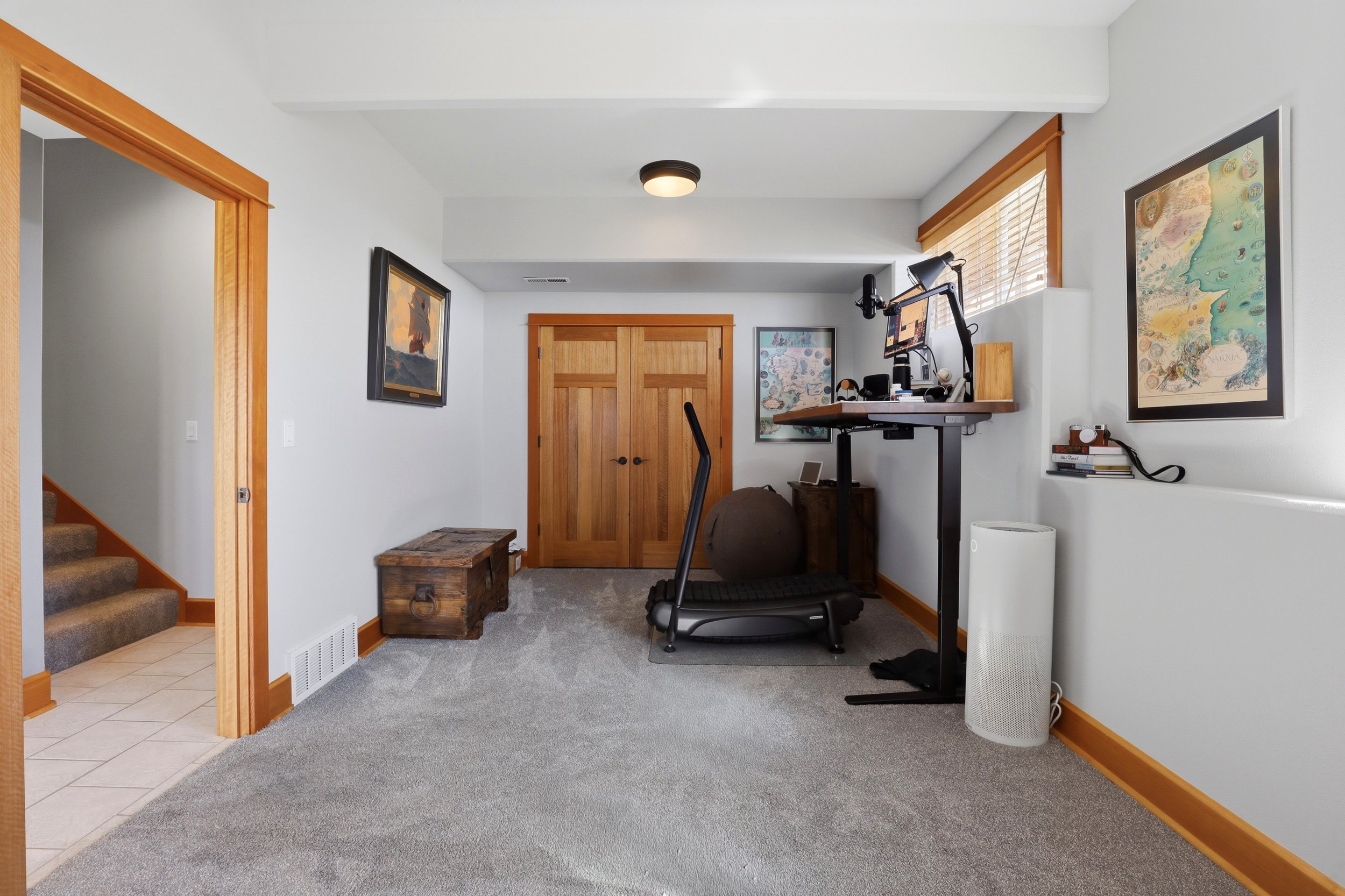 A home office space with a treadmill, a wooden chest, and artwork on the walls. Features include a ceiling light, a window with blinds, and a door with double wooden panels.