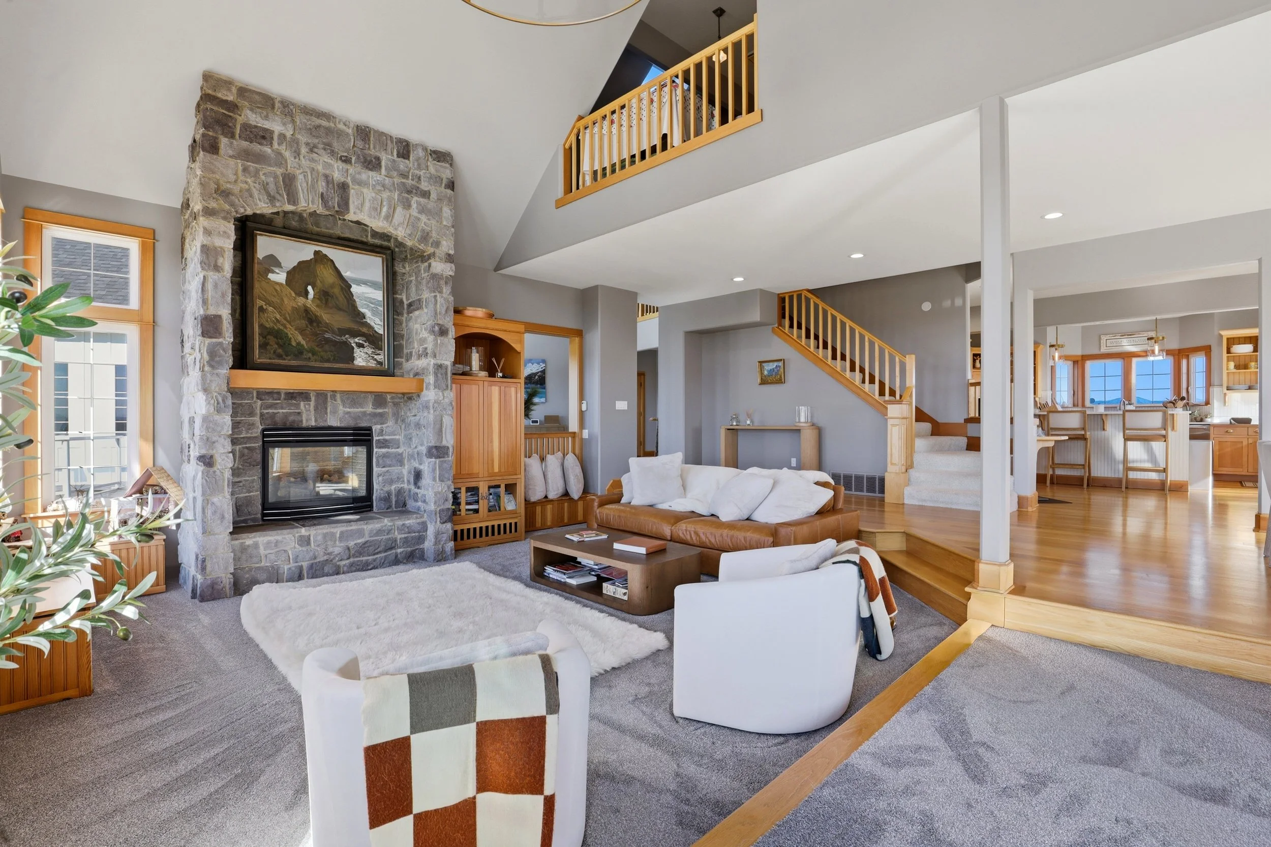 Spacious living room with a stone fireplace, wooden accents, and a vaulted ceiling. Features a white sofa, armchair, coffee table, and large windows.