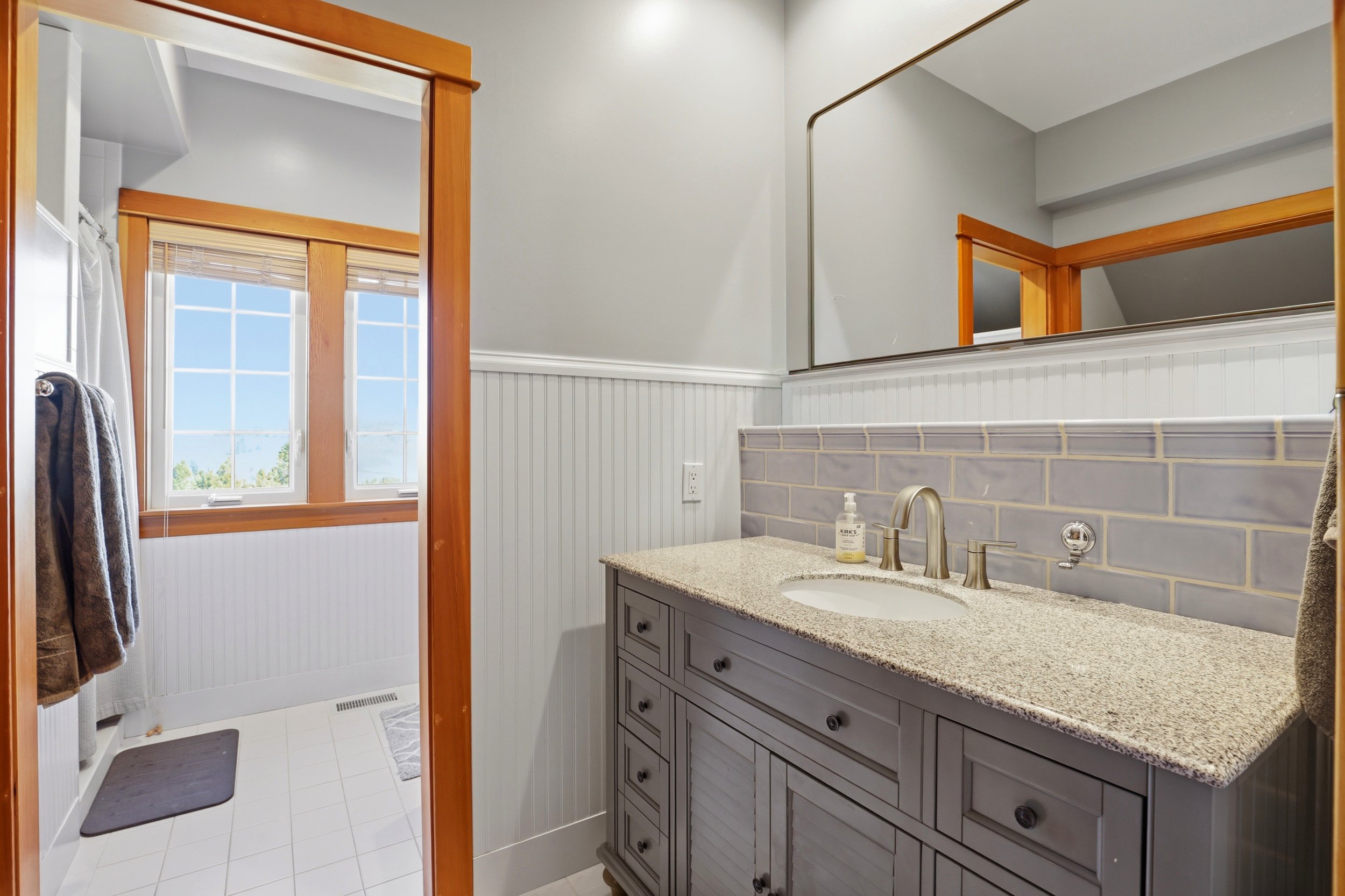 A bathroom with a beige granite countertop, gray wooden vanity cabinet, large mirror, white tiled backsplash, and a window with wooden trim and blinds. There are towels hanging on a towel bar near the shower and a soap dispenser on the counter.