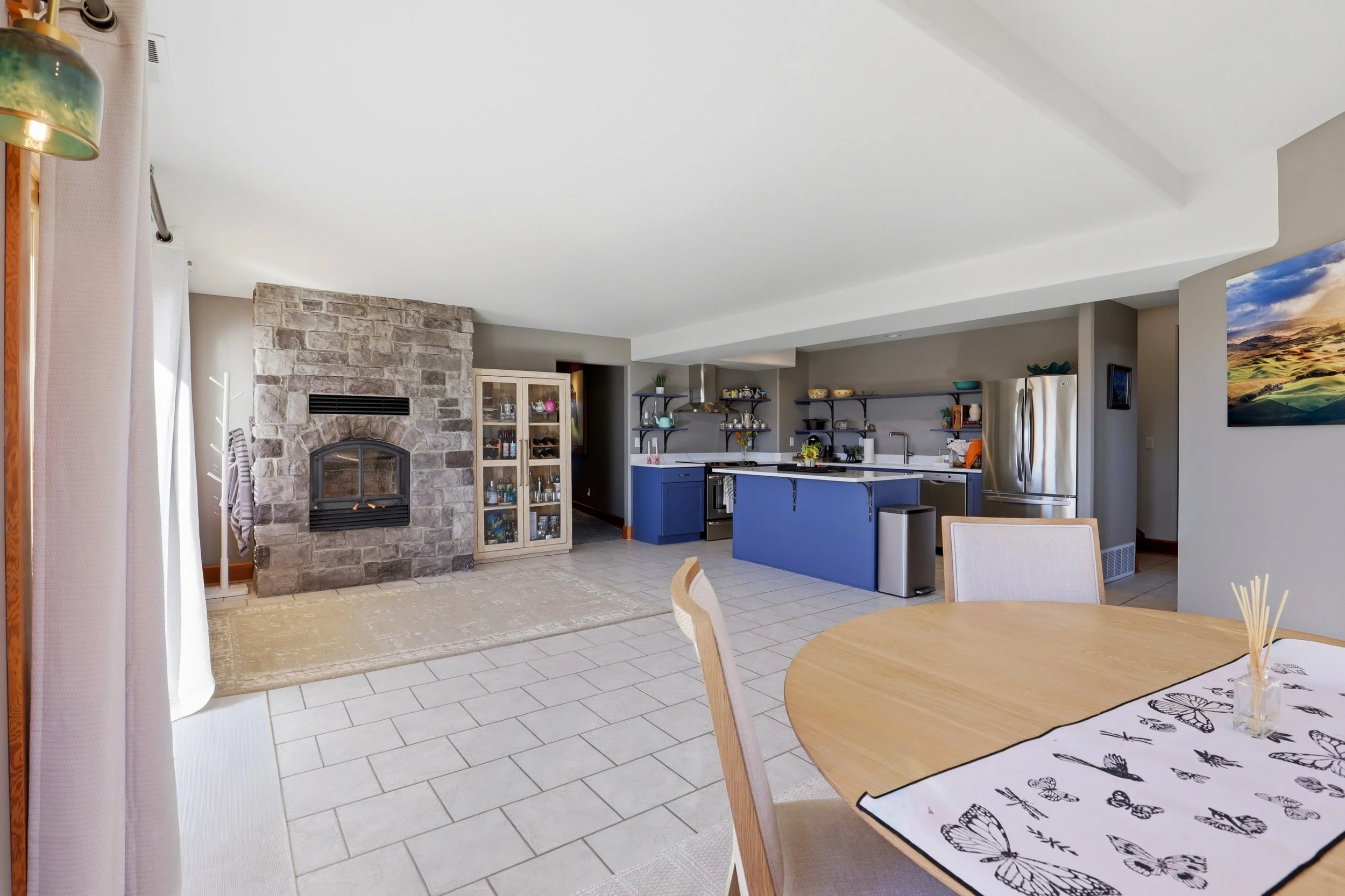 Open-concept living room and kitchen with stone fireplace, blue kitchen island, stainless steel refrigerator, open shelving, and a dining table with butterfly-themed tablecloth.