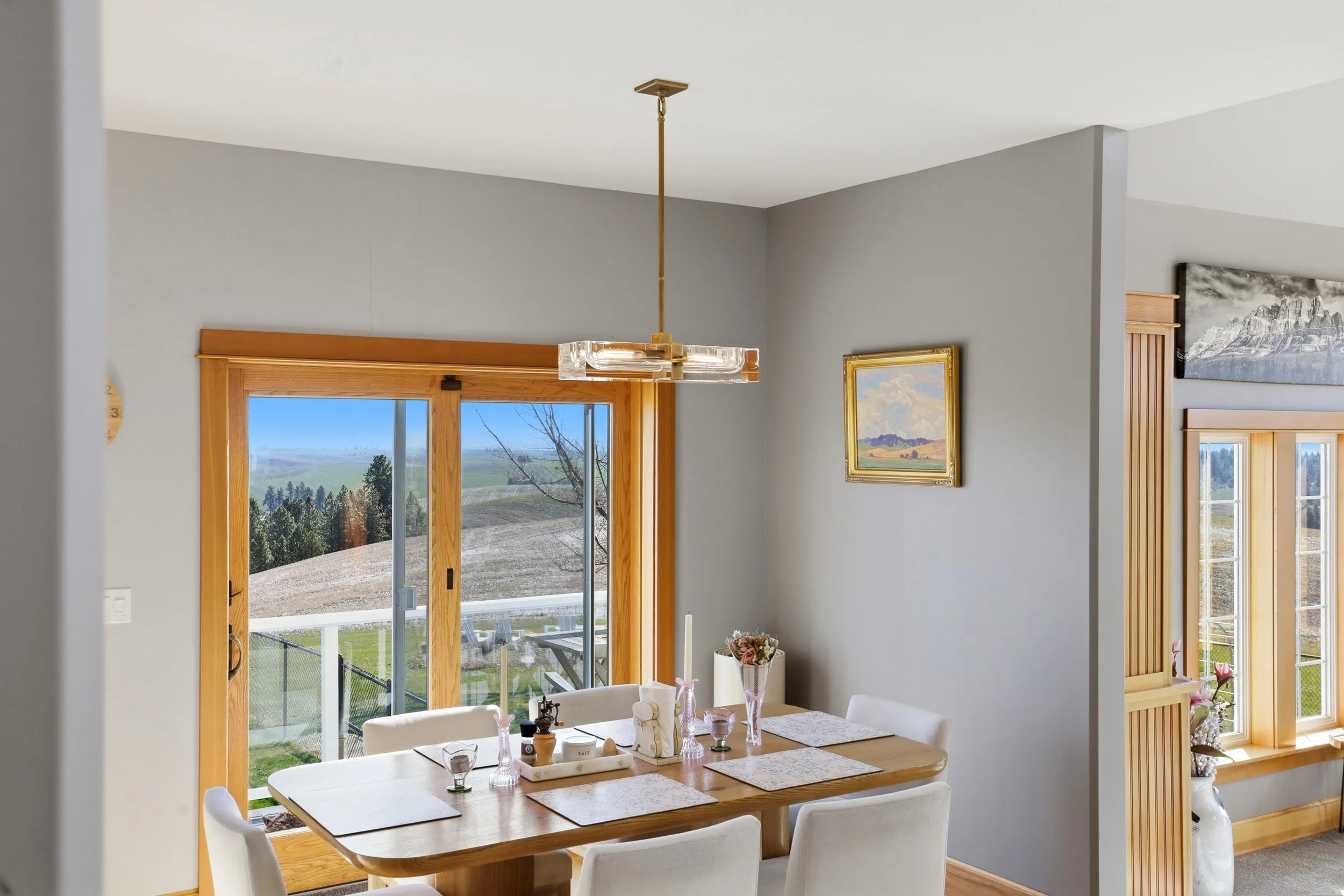 A dining room with a wooden table surrounded by white chairs, set with placemats, glassware, and centerpieces. Large glass sliding doors with wooden frames open to a view of a backyard with a fence, trees, and distant hills. The room has gray walls, 