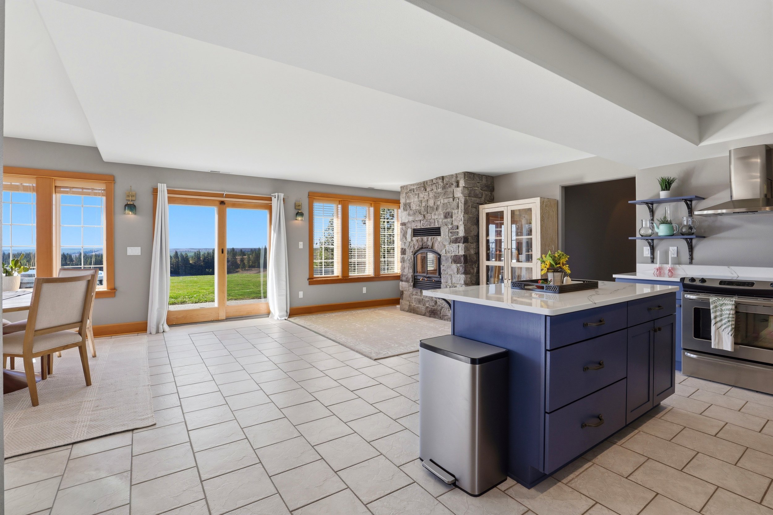 Open-concept living room and kitchen with large windows showing a green outdoor landscape; white tile floor, gray walls, stone fireplace, blue kitchen island, and dining area.