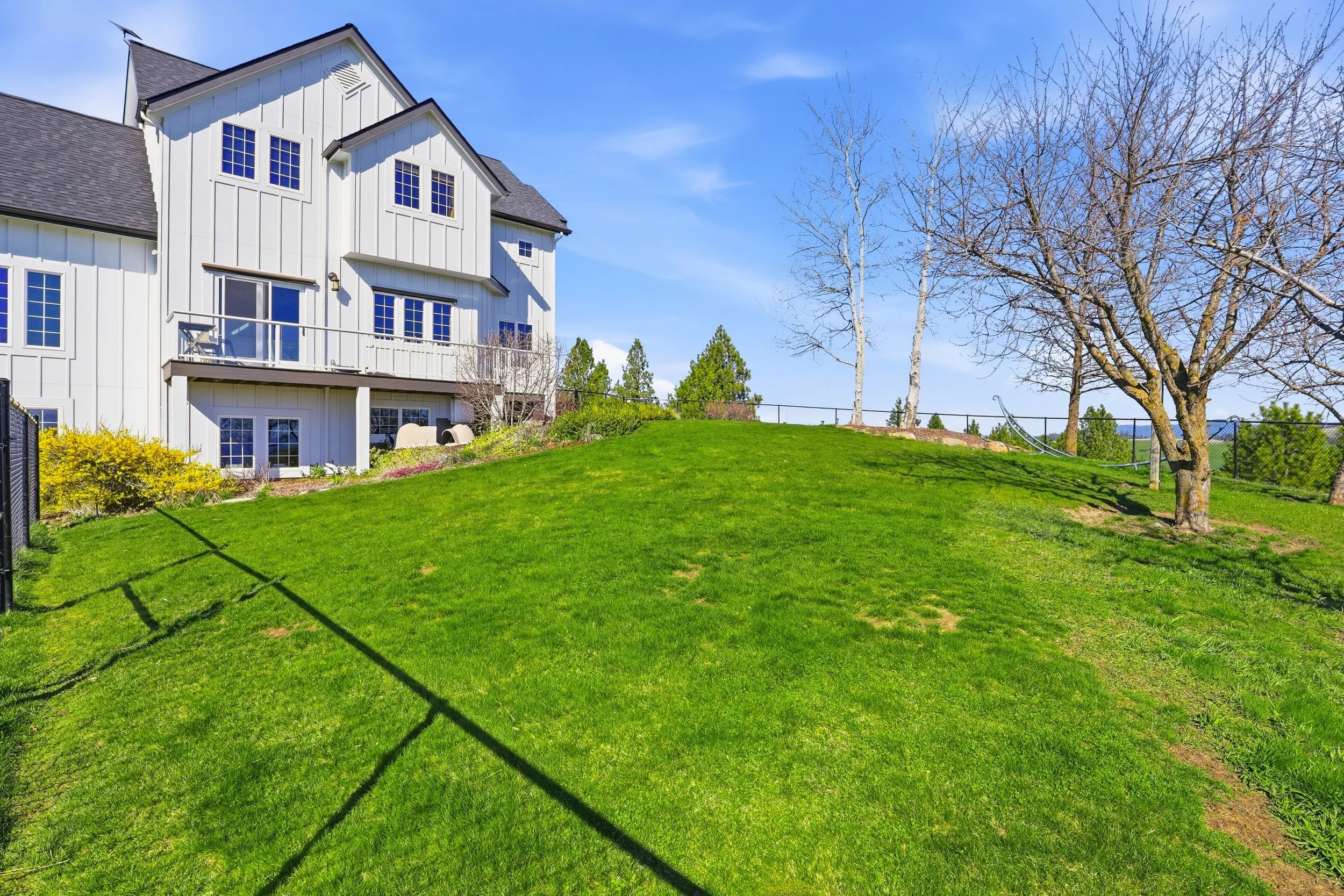 A white multi-story house with a gray roof, large windows, and a small balcony, surrounded by a green lawn and trees with some branches without leaves, under a blue sky.