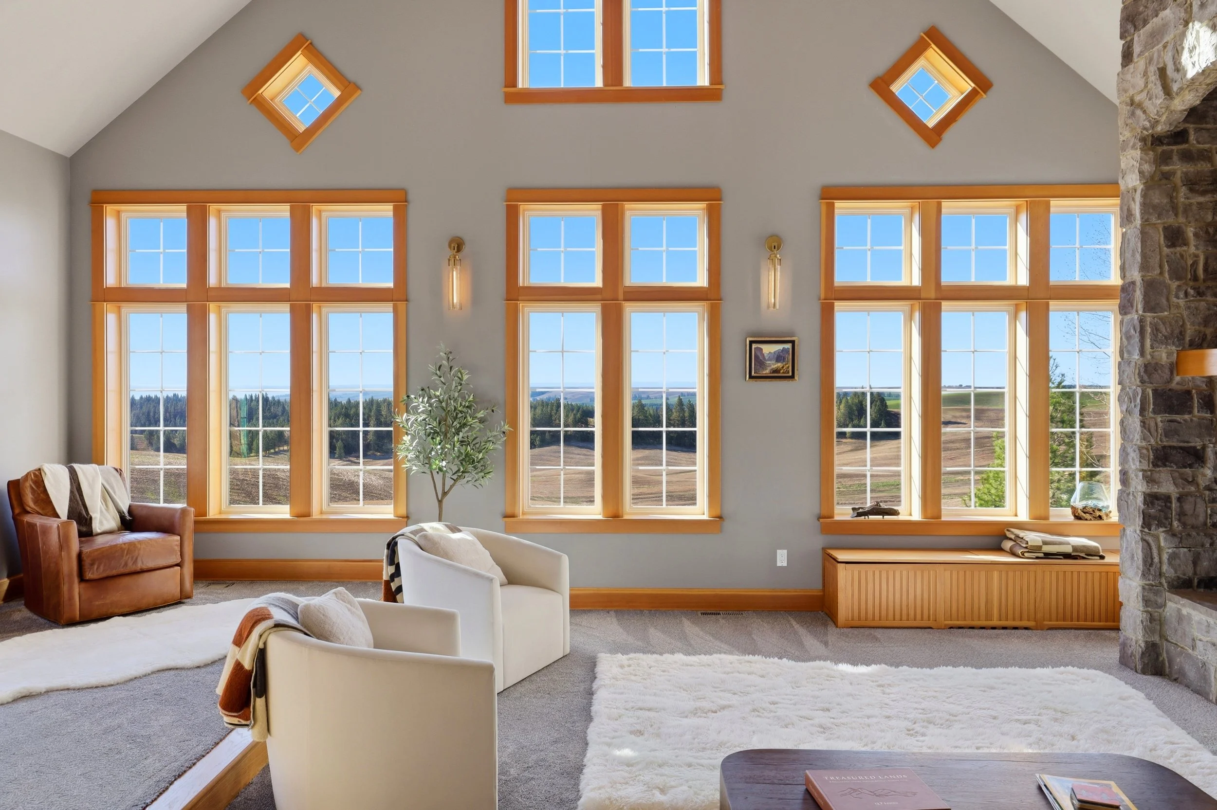 Living room with large multi-pane windows, a stone fireplace on the right, a leather armchair, a white armchair, a potted plant, and a painting on the wall.