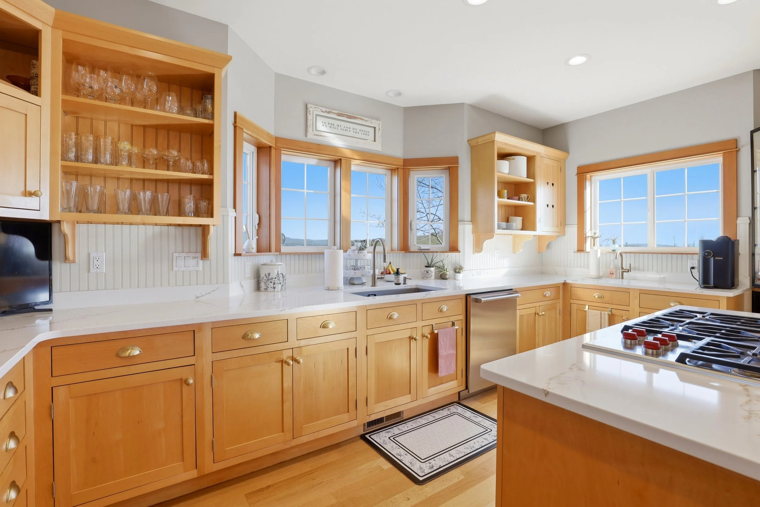 A bright kitchen with wooden cabinets, white countertops, and large windows showing a blue sky. The cabinets hold glasses and dishes, and a stove is visible in the foreground.