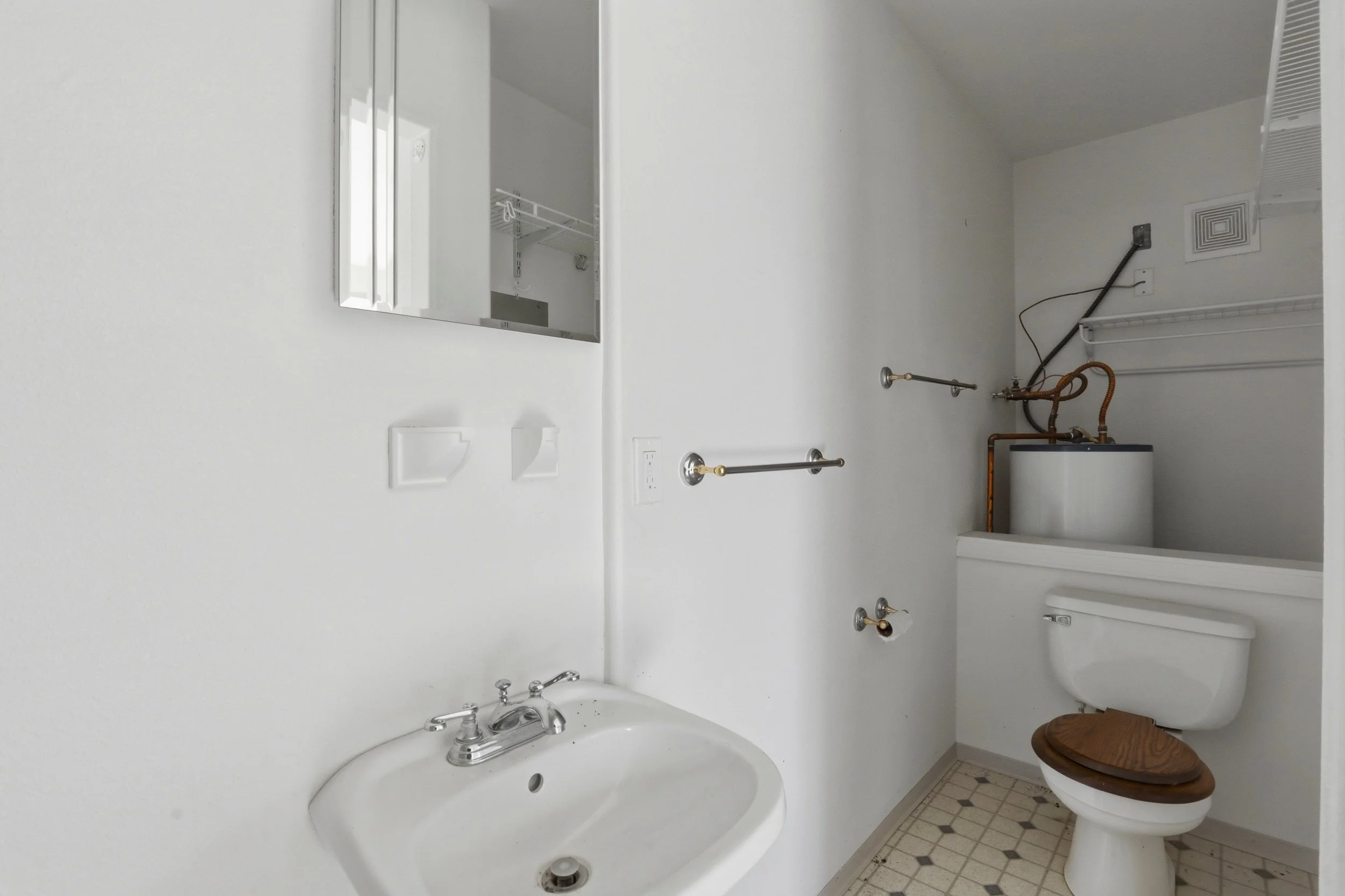 Empty bathroom with white walls, a toilet with a wooden seat, a white sink with a silver faucet, a mirror, and various plumbing fixtures, possibly indicating renovation or repair.
