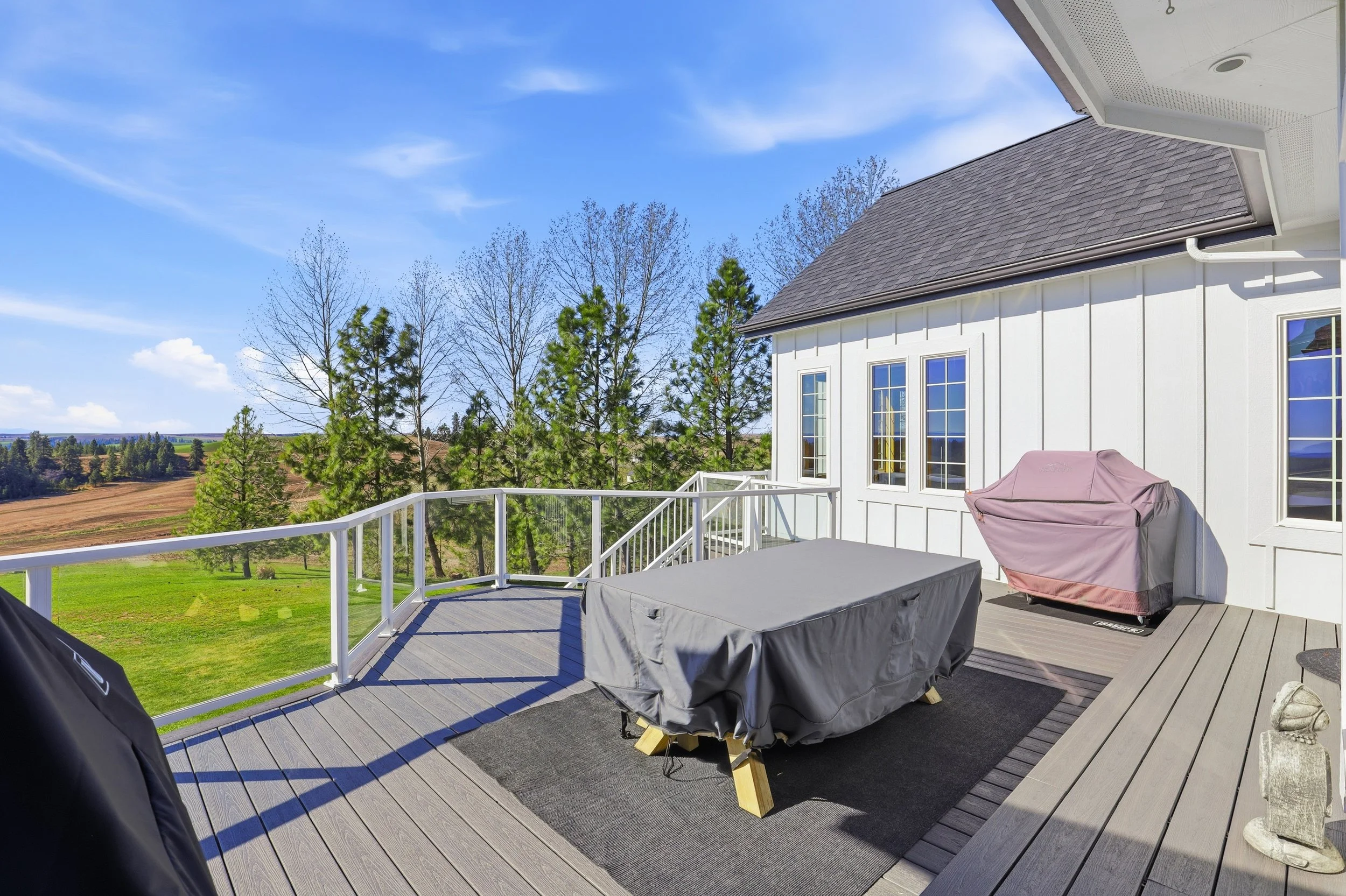 View of a suburban backyard deck with outdoor furniture and a grill, overlooking a green lawn and trees under a blue sky.