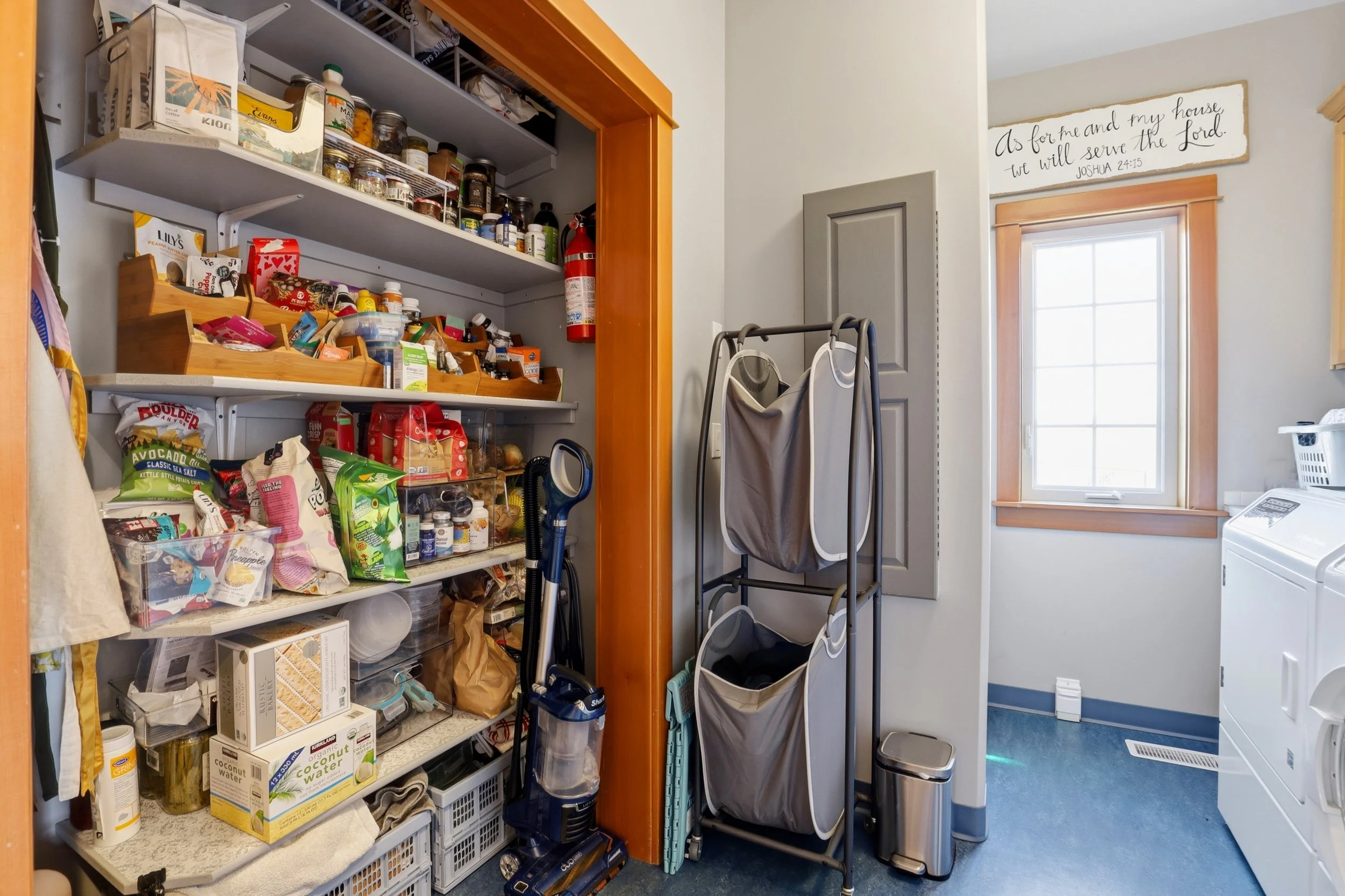 Closet and laundry room with shelves filled with food and household supplies, laundry hamper, and a washing machine near a window with a sign above it reading, "As for me and my house we will serve the Lord. Joshua 24:15."