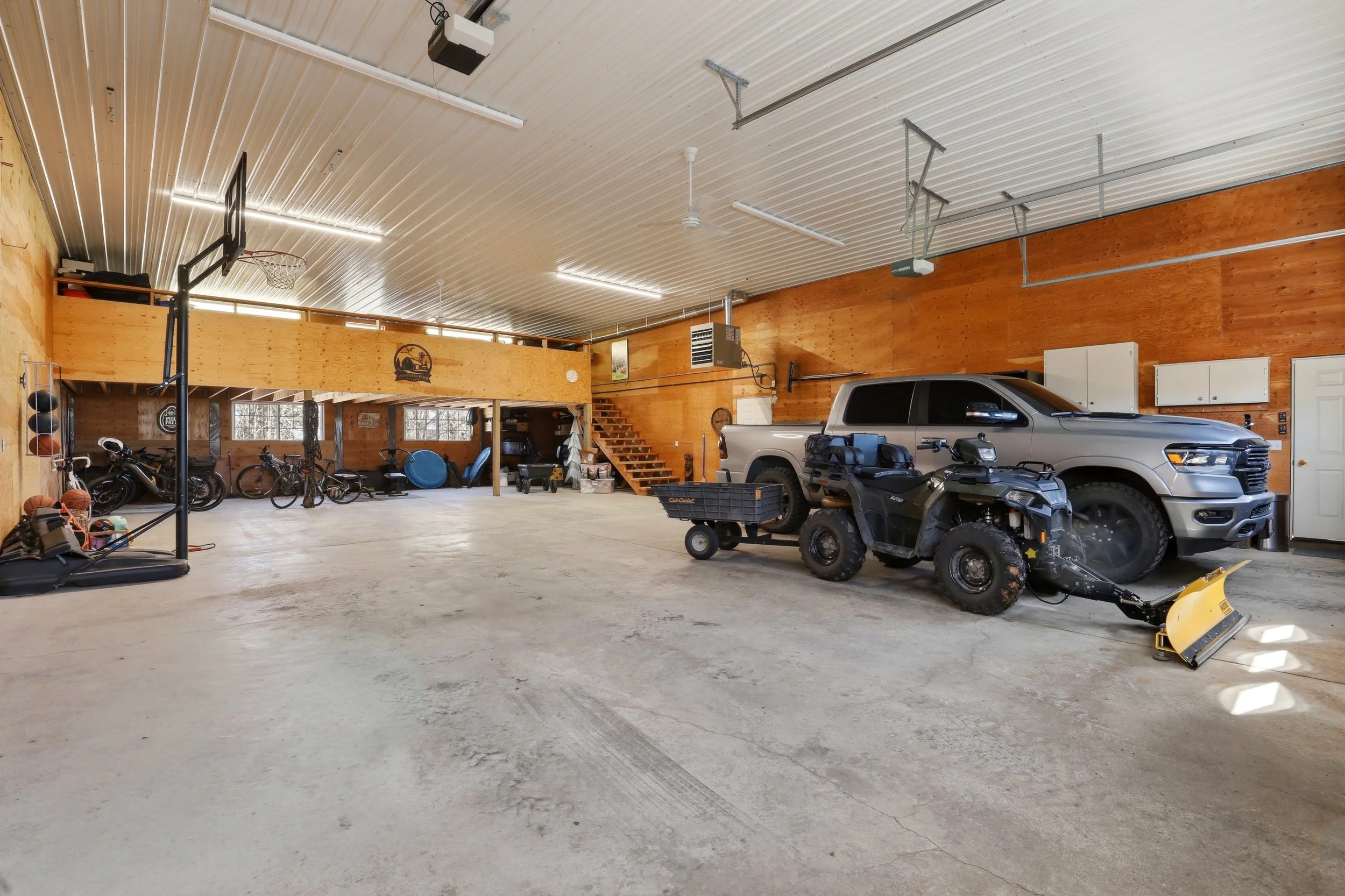 A spacious garage with a high metal ceiling, wooden walls, and concrete floor, containing a black trampoline, motorcycles, bicycles, a pickup truck, an ATV, and various tools and equipment.