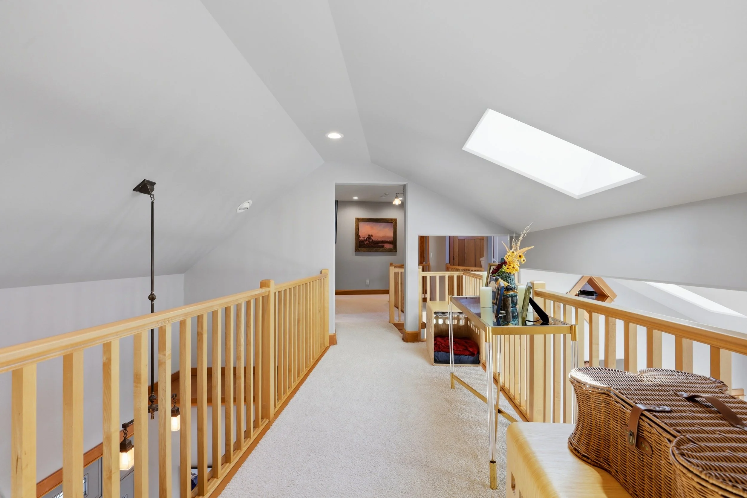 A spacious, bright attic living area with white sloped ceiling, skylight, wooden railing, a table with a sunflower arrangement, and wicker chair.